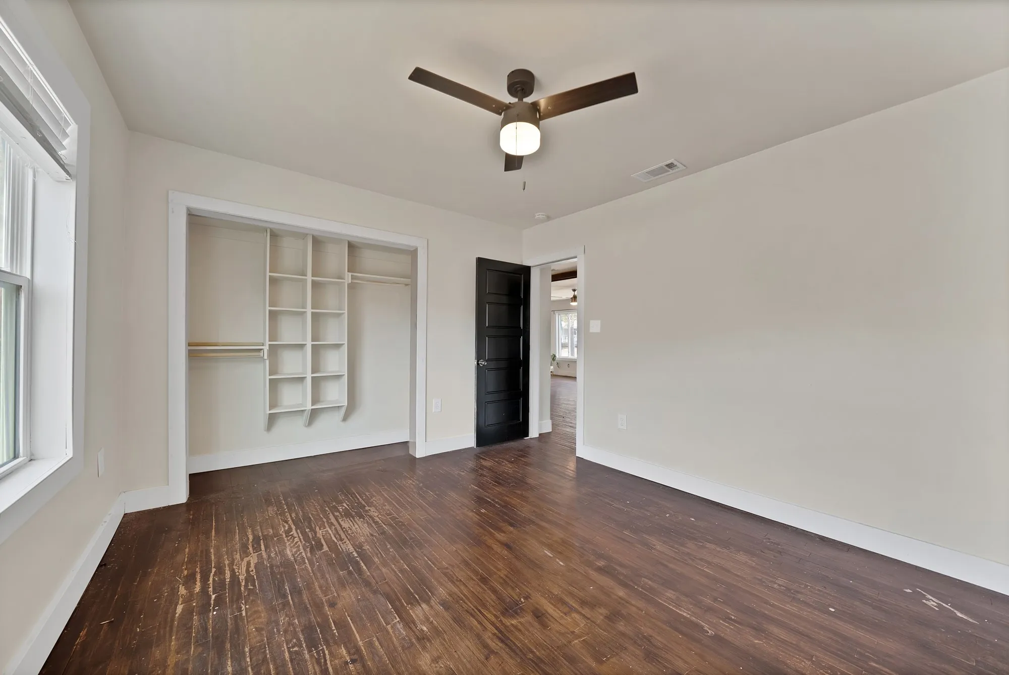 Bedroom: Reach-in closet with built-in shelving organizer and hanging rods, ceiling fan with light, and window with blinds. Black paneled interior door and hardwood floors.