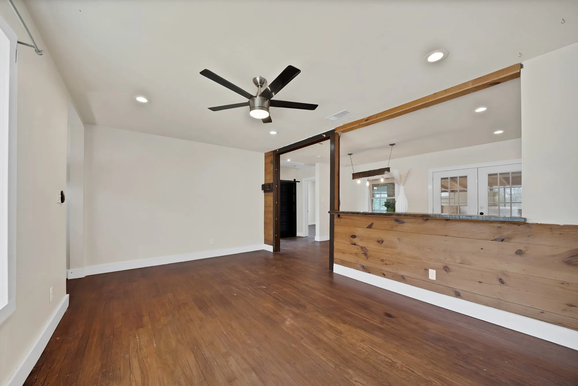 Living room: Knotty pine plank breakfast bar with pass-through to the dining area, exposed wood beam header, and dark timber posts. Ceiling fan with light, recessed lighting, hardwood floors, and sliding barn door.