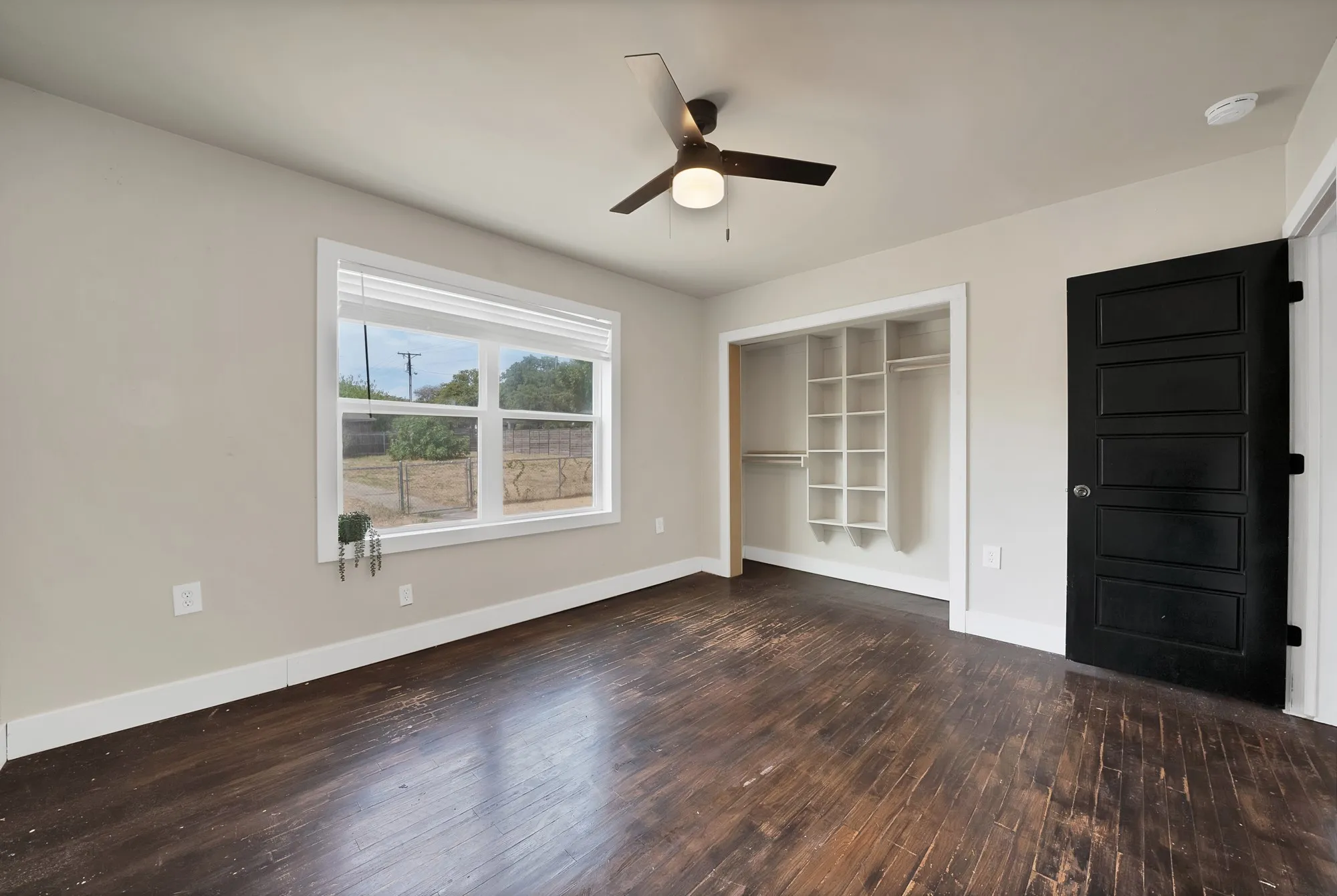 Bedroom: Large window with blinds overlooking the backyard, ceiling fan with light, and hardwood floors. Reach-in closet with built-in cubby shelving and hanging rods, and black paneled interior door.