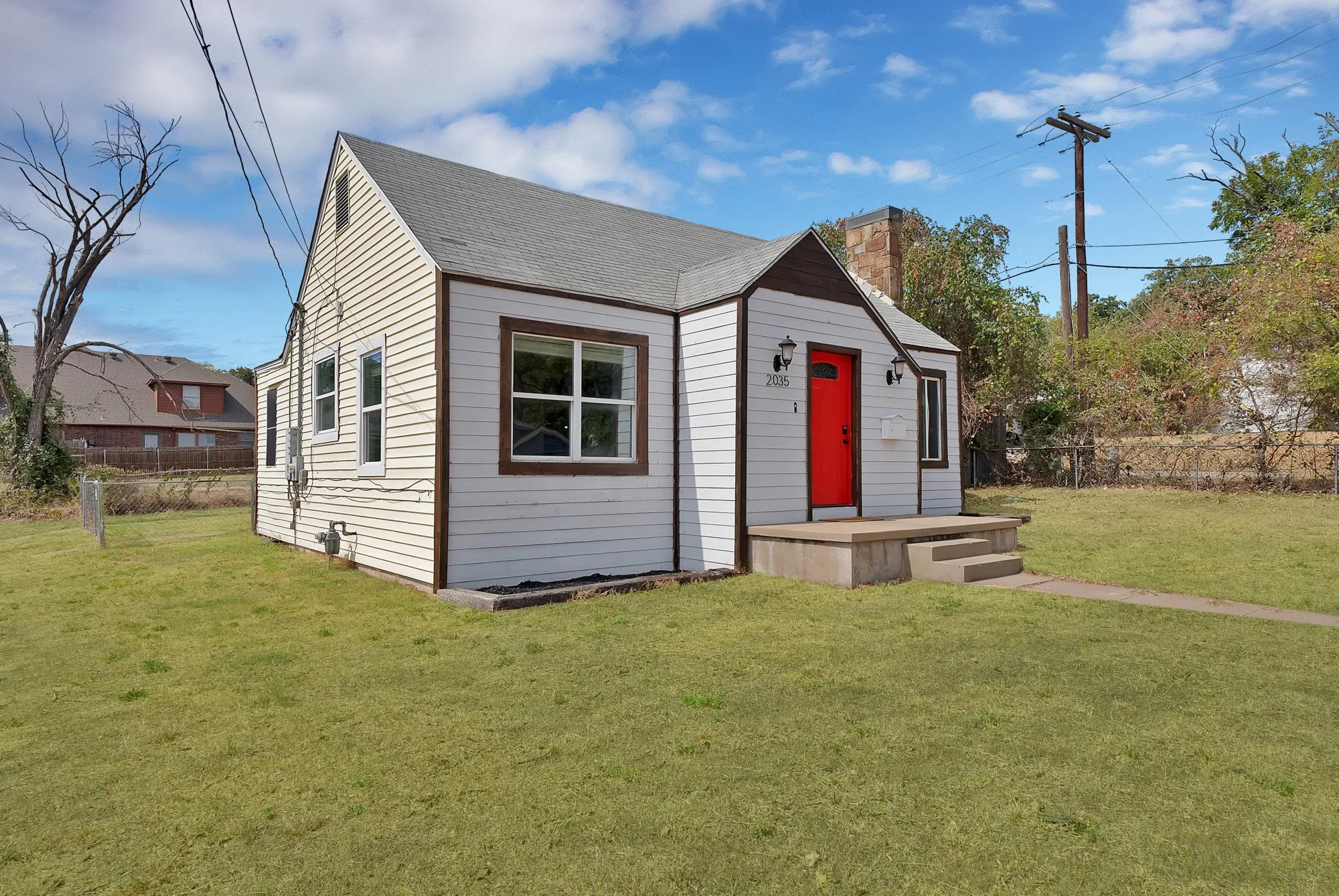 Front exterior: Stone chimney, oversized picture window, and red front door with lantern-style sconces. Central air conditioning unit, landscaped bed borders, and wood privacy fence.