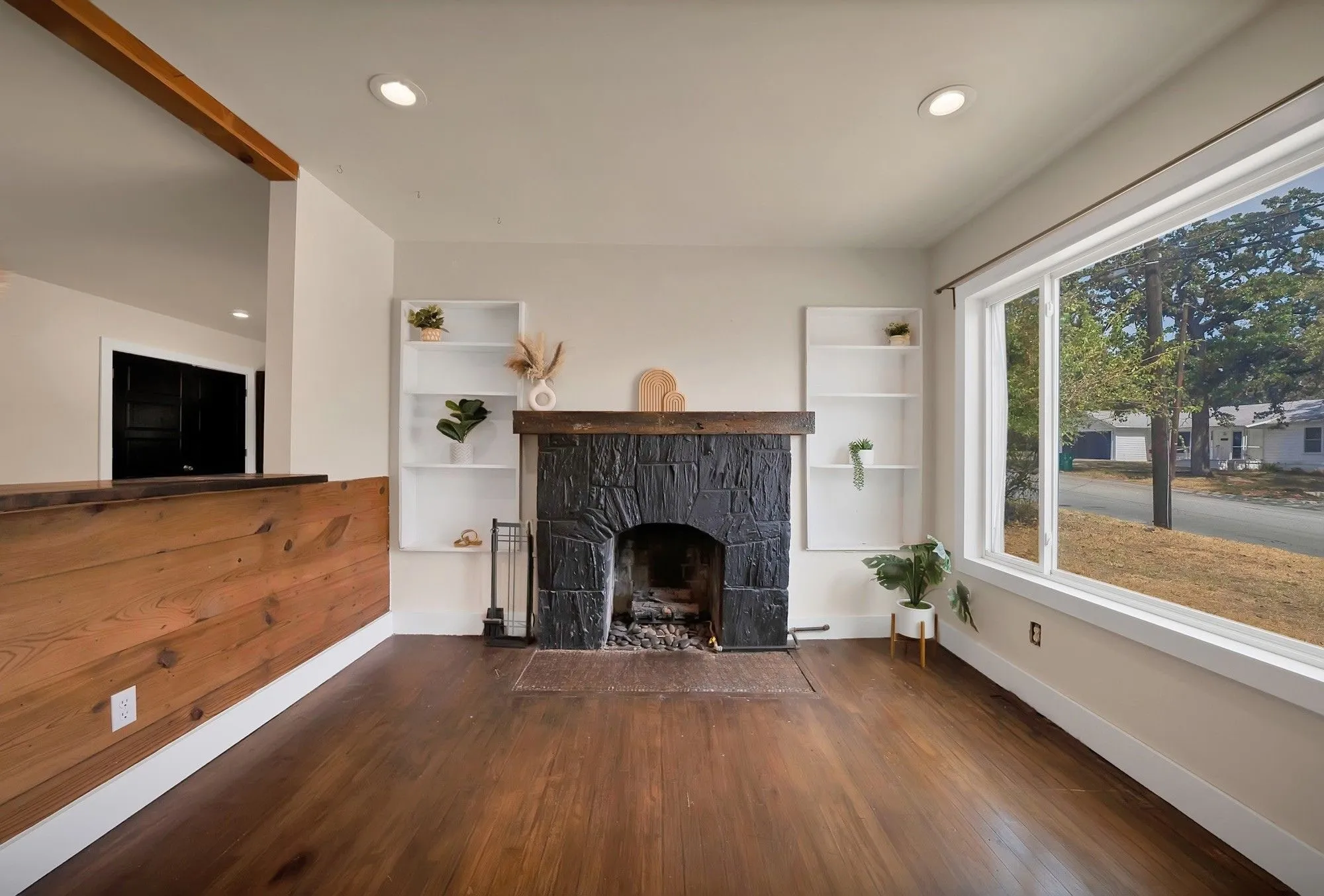 Living room: Black painted stone fireplace with arched firebox, reclaimed wood beam mantel, and flanking built-in bookshelves. Oversized picture window, recessed lighting, hardwood floors, and exposed wood beam opening to the dining area.