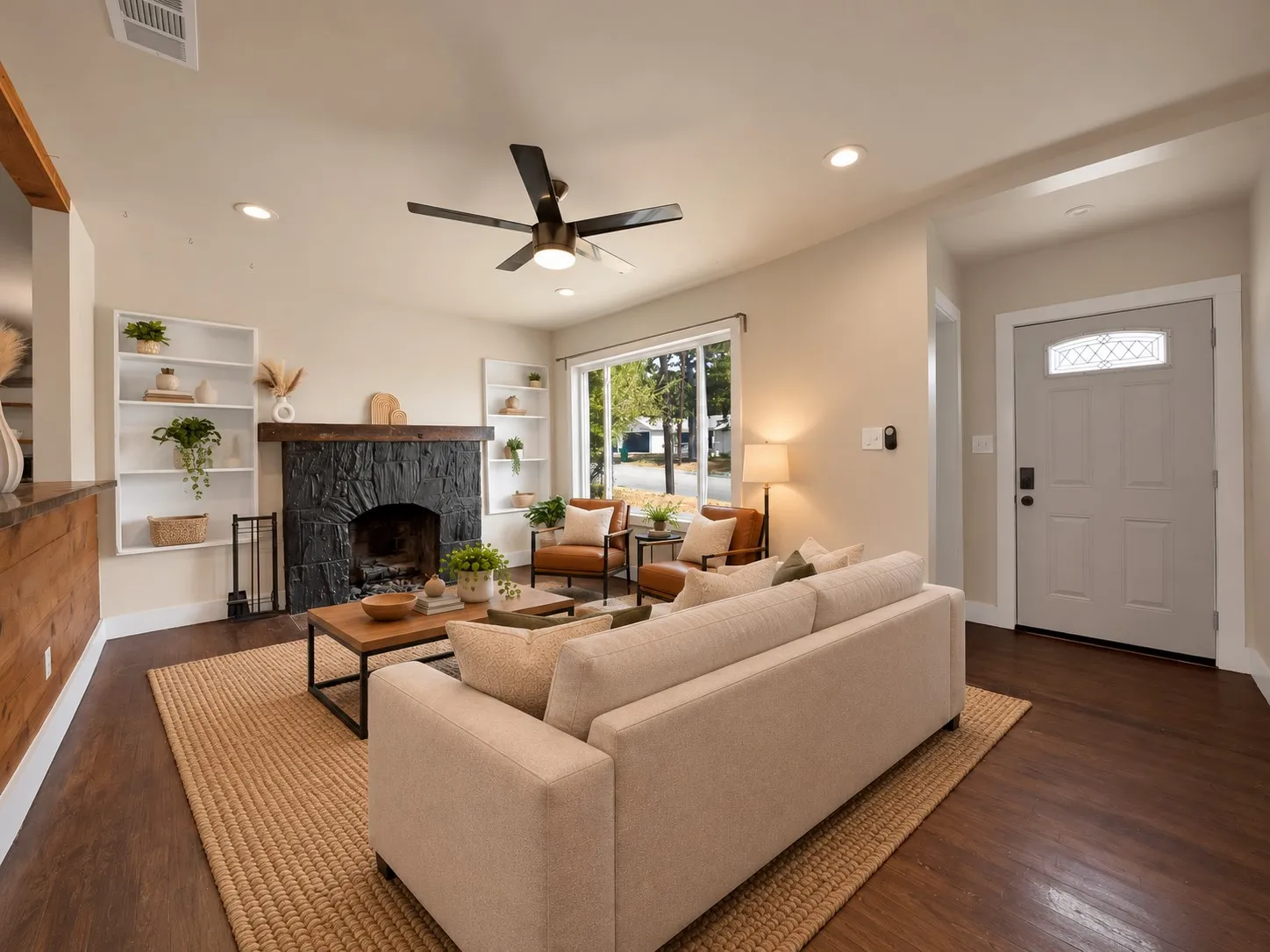 virtually staged Living room: Black painted stone fireplace with reclaimed wood beam mantel and flanking built-in bookshelves. Oversized picture window, ceiling fan with light, recessed lighting, hardwood floors, and front entry door with keypad smart lock.