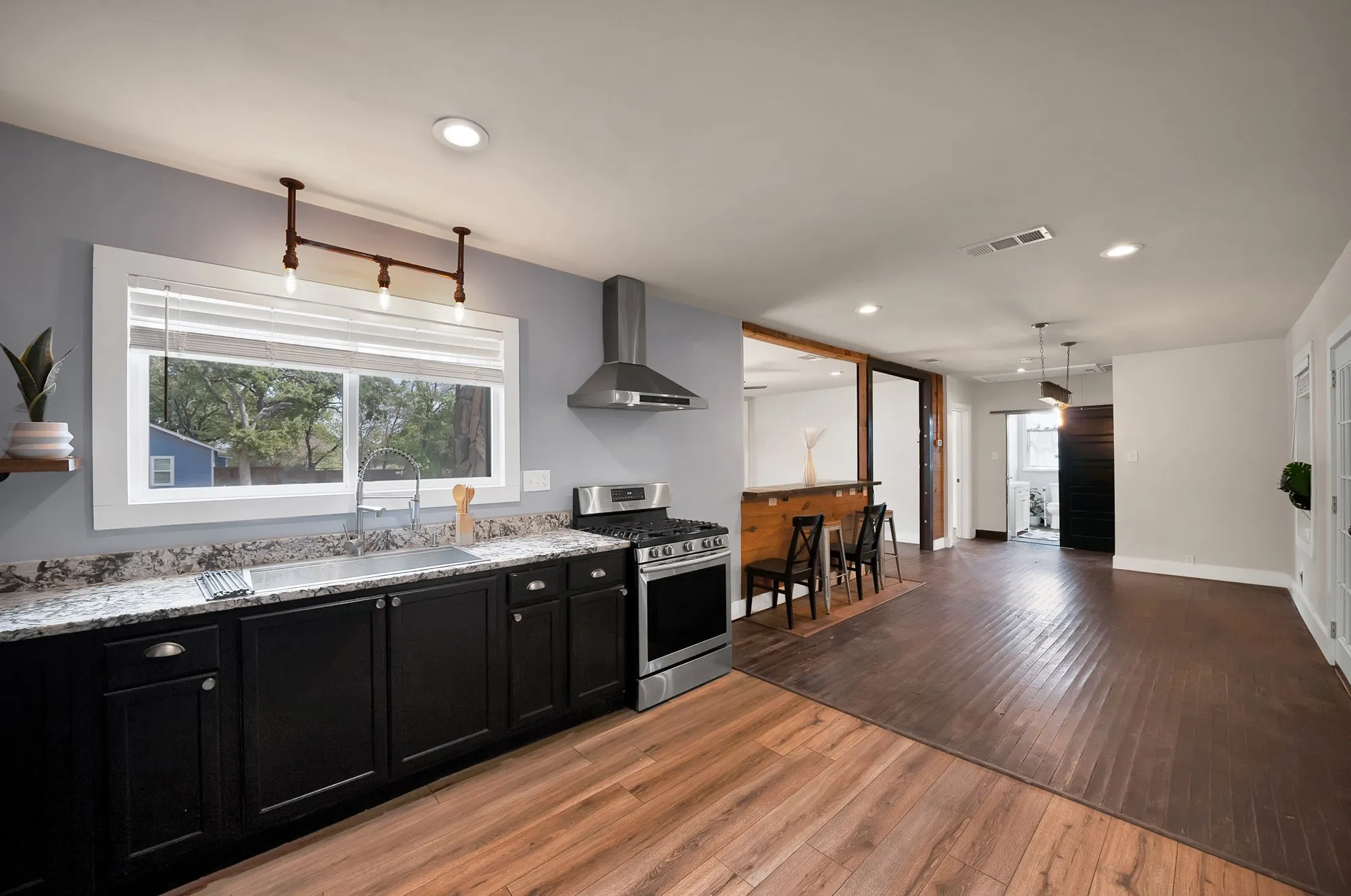 Kitchen: Black shaker cabinetry, granite countertops, and window over the sink with industrial pipe pendant lighting. Open floor plan flows past the knotty pine breakfast bar into the dining area, with wood-look plank flooring transitioning to hardwood floors.