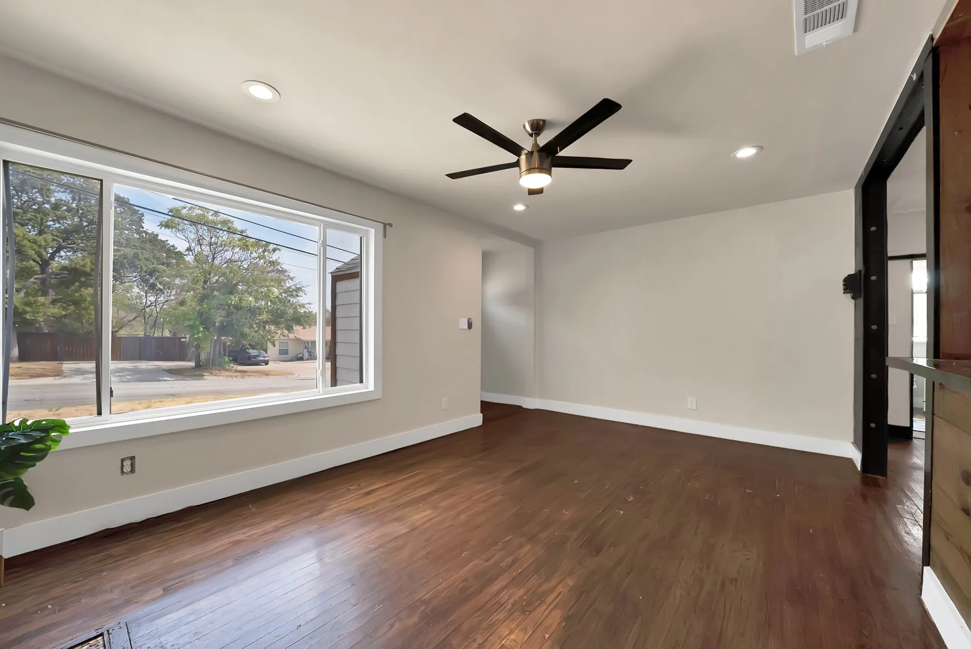Living room: Oversized picture window, ceiling fan with light, recessed lighting, and hardwood floors. Wood plank and dark timber framed opening connects to the dining area.