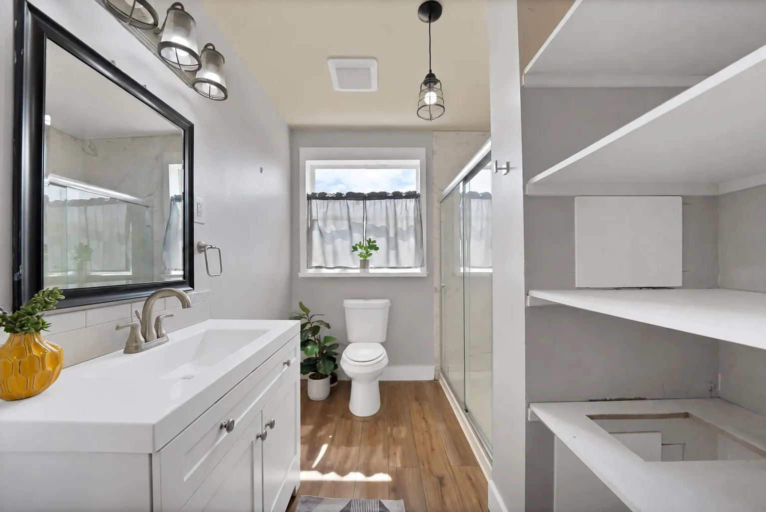 Bathroom: White shaker vanity with brushed nickel faucet, subway tile backsplash, and industrial cage pendant lighting. Walk-in shower with sliding glass doors, window, and built-in linen shelving for storage.