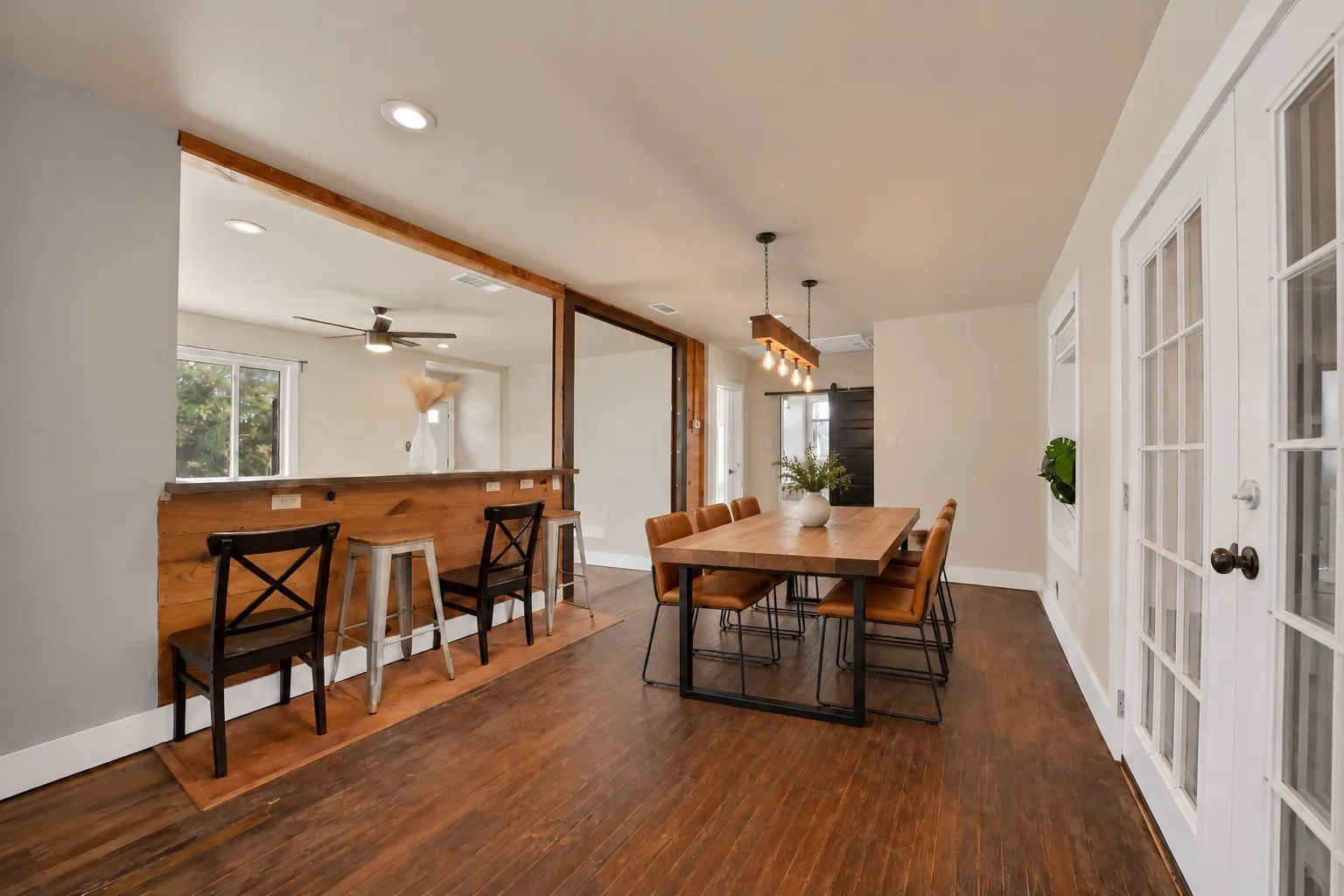 virtually staged Dining area: Wood beam chandelier with Edison bulb pendants, French doors, and hardwood floors. Knotty pine plank breakfast bar with seating, pass-through to the living room, and sliding barn door.