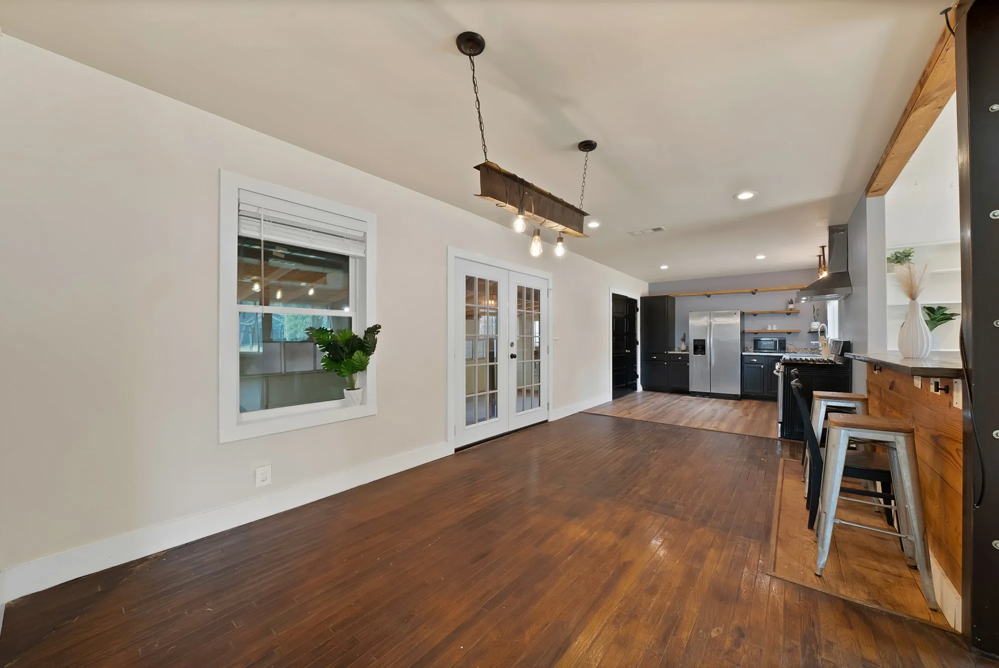 Dining area: Industrial steel I-beam chandelier with Edison bulb pendants, French doors to the covered back patio, and hardwood floors. Open floor plan flows into the kitchen and knotty pine breakfast bar with seating.