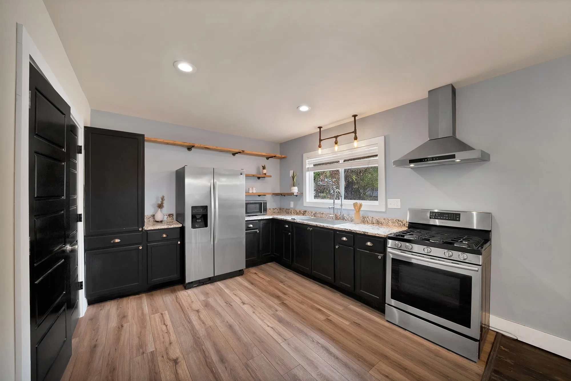 Kitchen: L-shaped layout with granite countertops, stainless steel sink, and pull-down faucet under a window. Black shaker cabinetry with cup pulls, tall storage cabinet, open wood floating shelves, recessed lighting, and wood-look plank flooring.