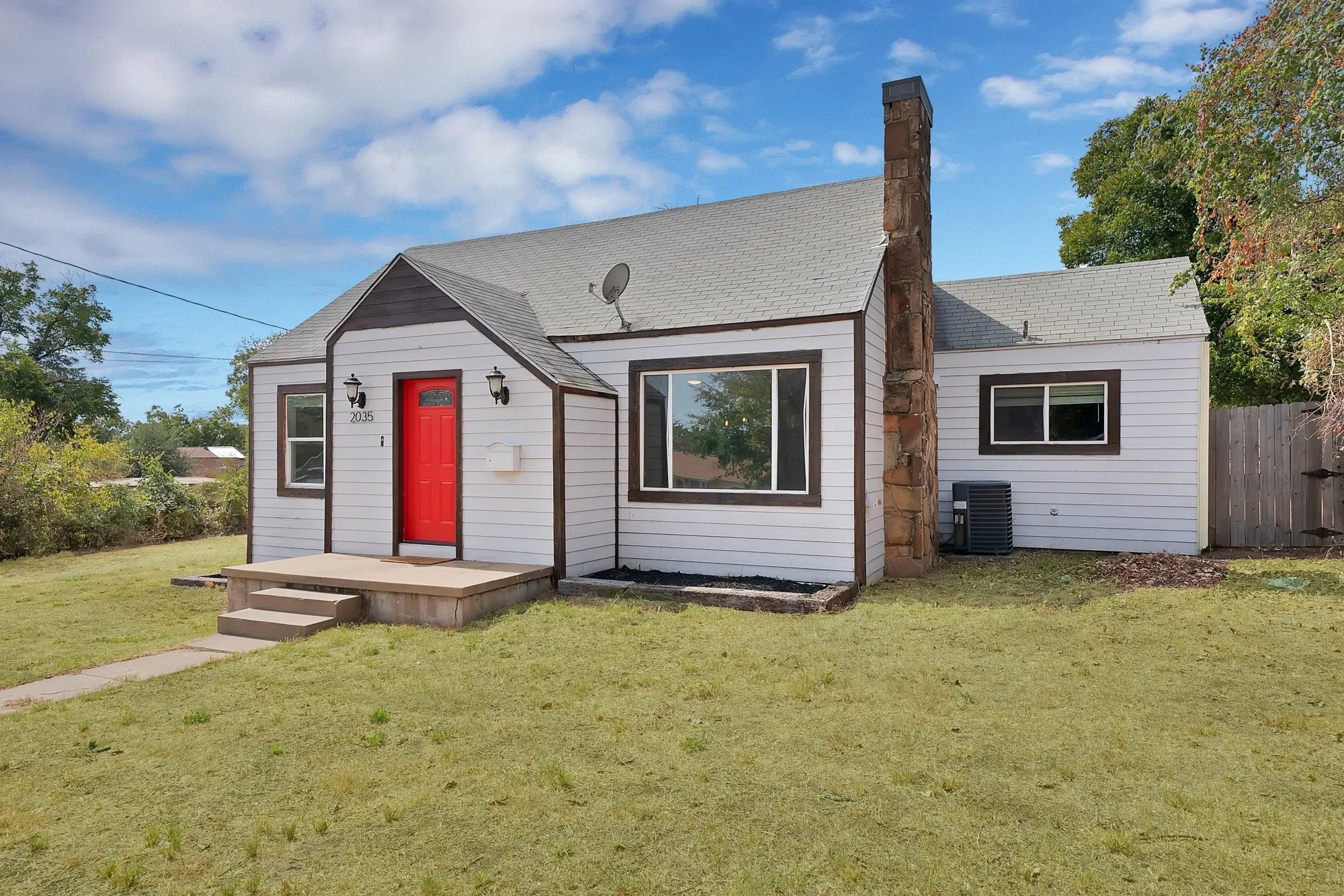 Front exterior: Stone chimney, oversized picture window, and red front door with lantern-style sconces. Central air conditioning unit, landscaped bed borders, and wood privacy fence.