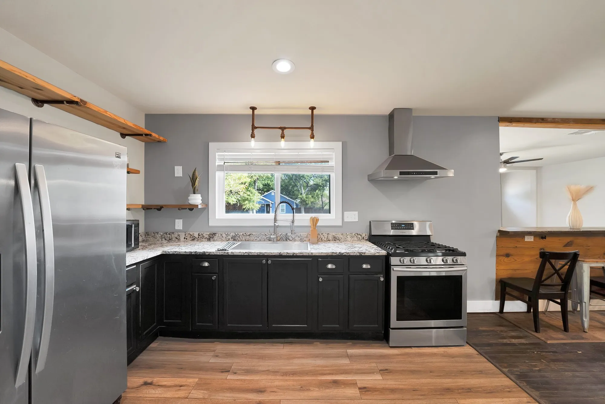 Kitchen: Black shaker cabinetry, granite countertops, and stainless steel appliances with Samsung gas range, side-by-side refrigerator, and chimney vent hood. Open wood floating shelves on pipe brackets, industrial pipe pendant with Edison bulbs, and wood-look plank flooring.