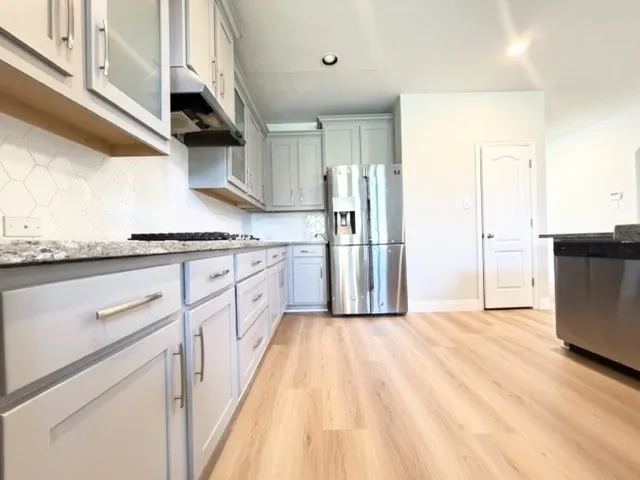 Light wood-finish flooring, light gray cabinetry with brushed nickel hardware, stainless steel appliances, speckled stone countertops, and a white hexagonal tile backsplash