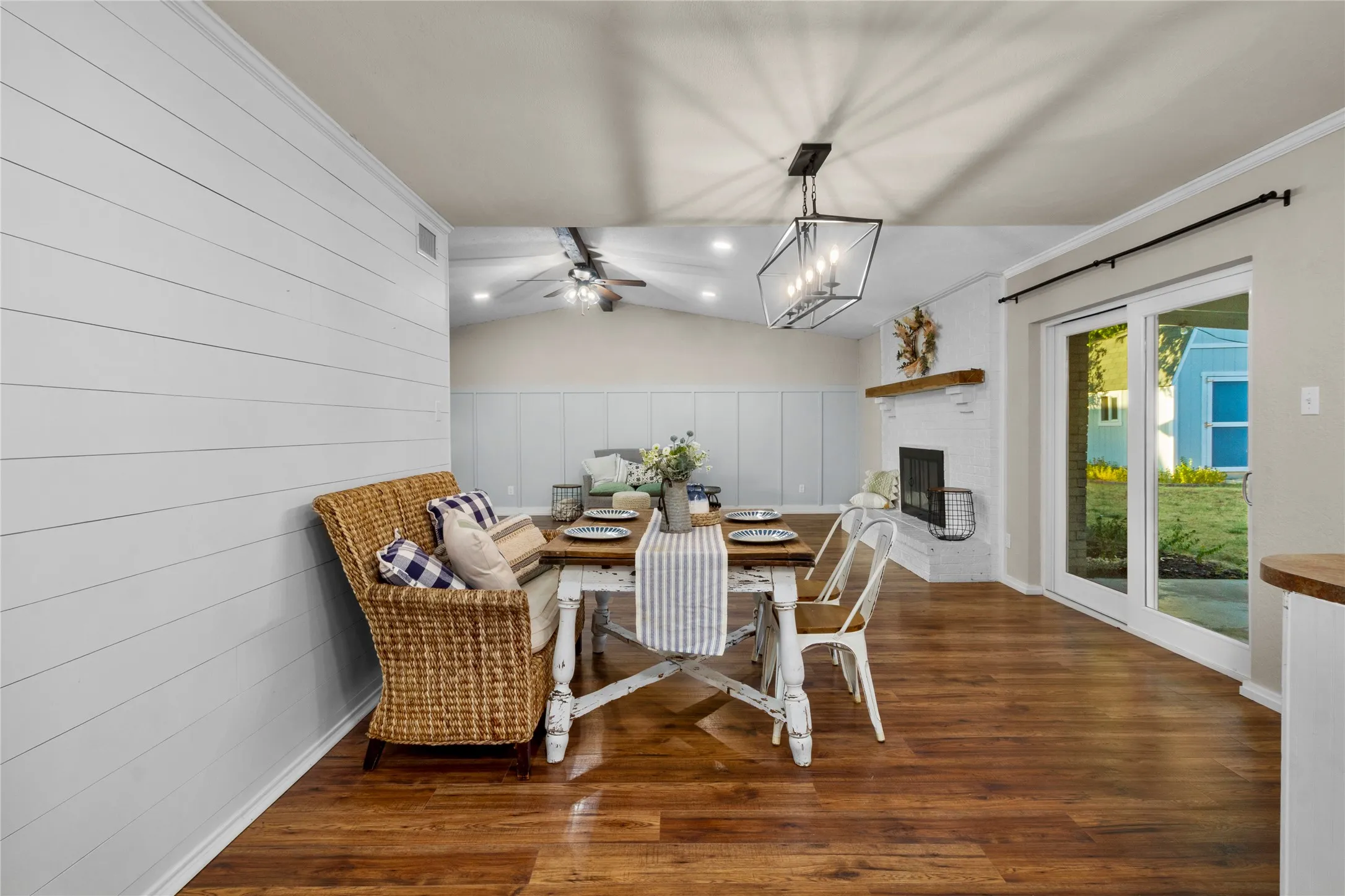 Warm wood-finish flooring, white shiplap accent wall, modern linear chandelier, white brick fireplace with a wood mantel, and sliding glass patio doors