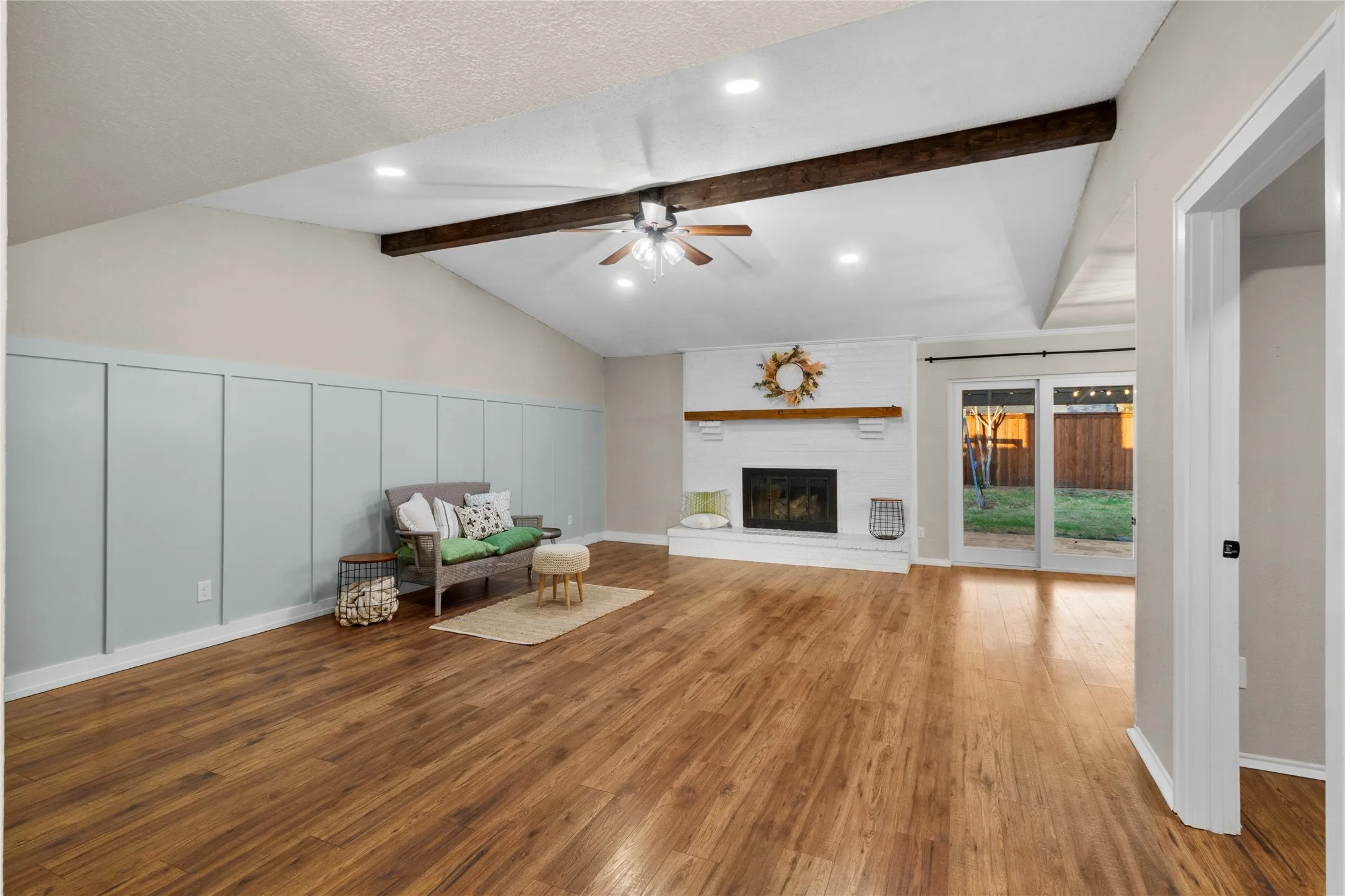 Vaulted ceiling with exposed beam, recessed lighting, wood-finish flooring, white brick fireplace with wood mantel, and board and batten wall accent