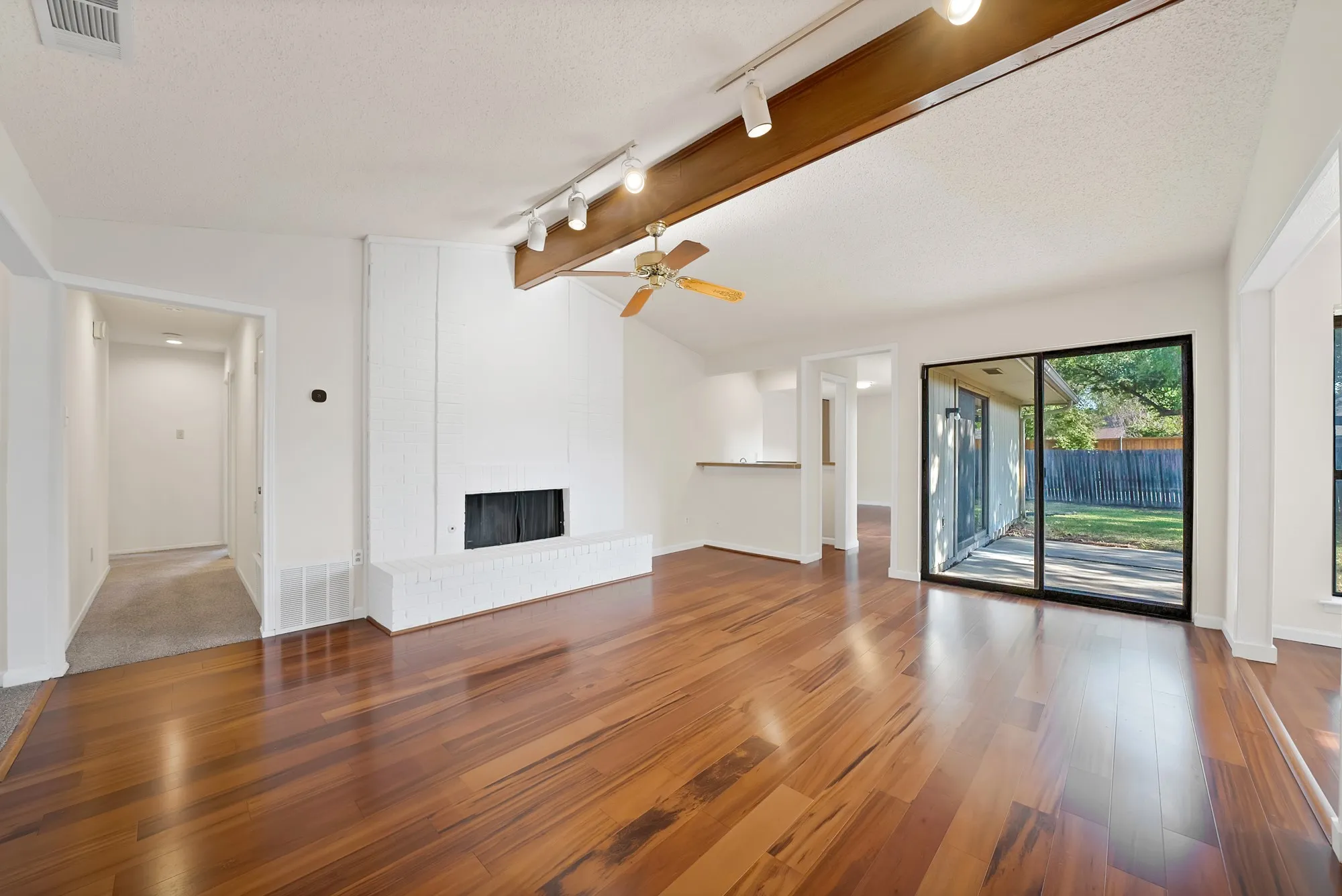 Vaulted ceiling with wood beam accent, wood-finish flooring, white brick fireplace, sliding glass door, and a ceiling fan