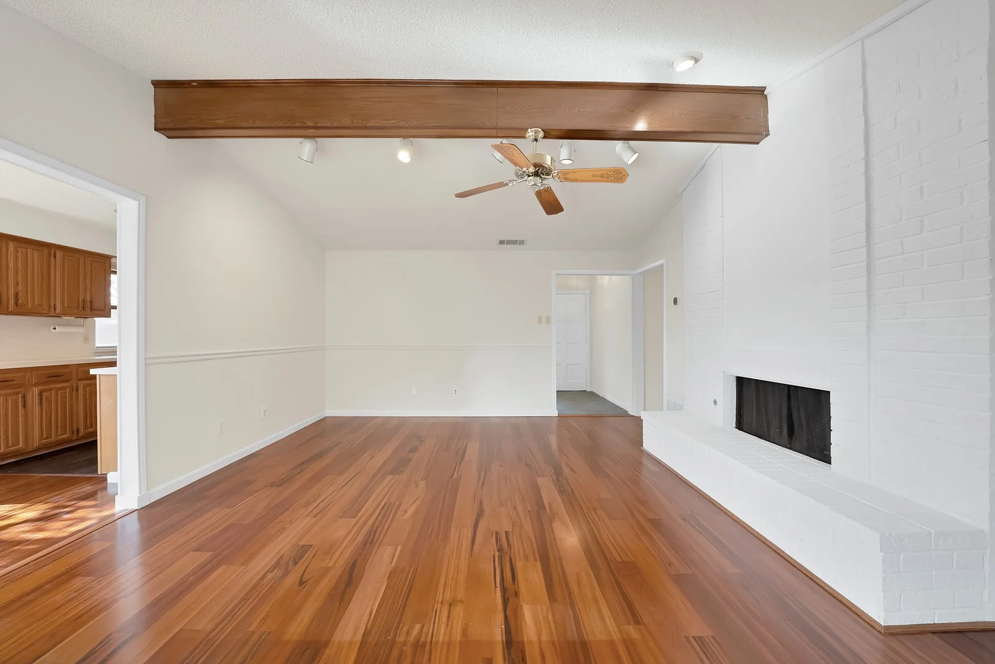 Vaulted ceiling with exposed wood beam, wood-finish flooring, painted brick fireplace, track lighting, and a ceiling fan