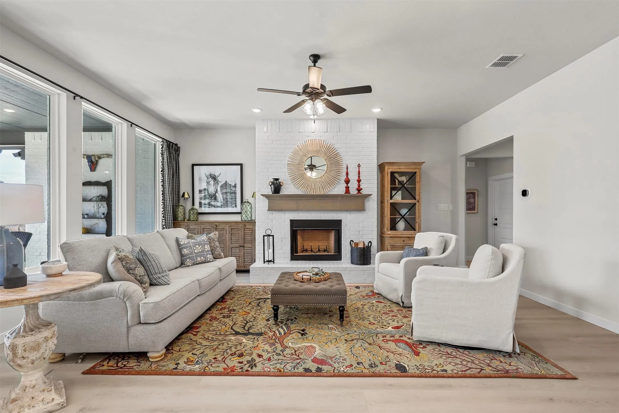 Light wood-finish flooring, white brick fireplace with a wood mantel, ceiling fan with integrated lighting, recessed lighting, and large windows