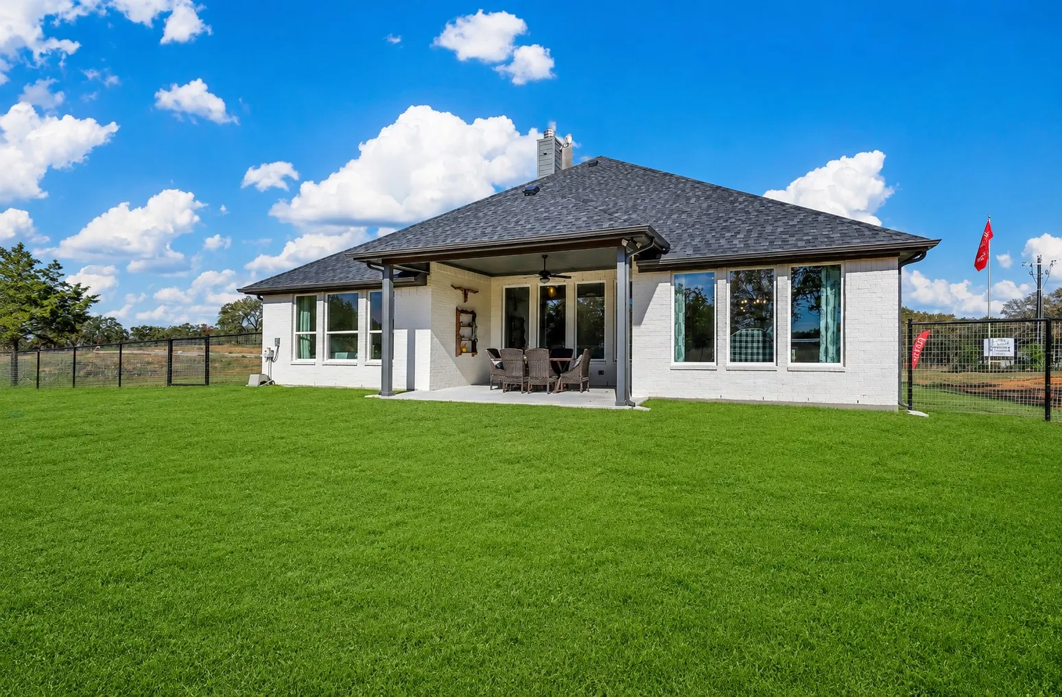 Expansive green lawn with a covered patio featuring an outdoor ceiling fan, white brick exterior, and a dark shingle roof