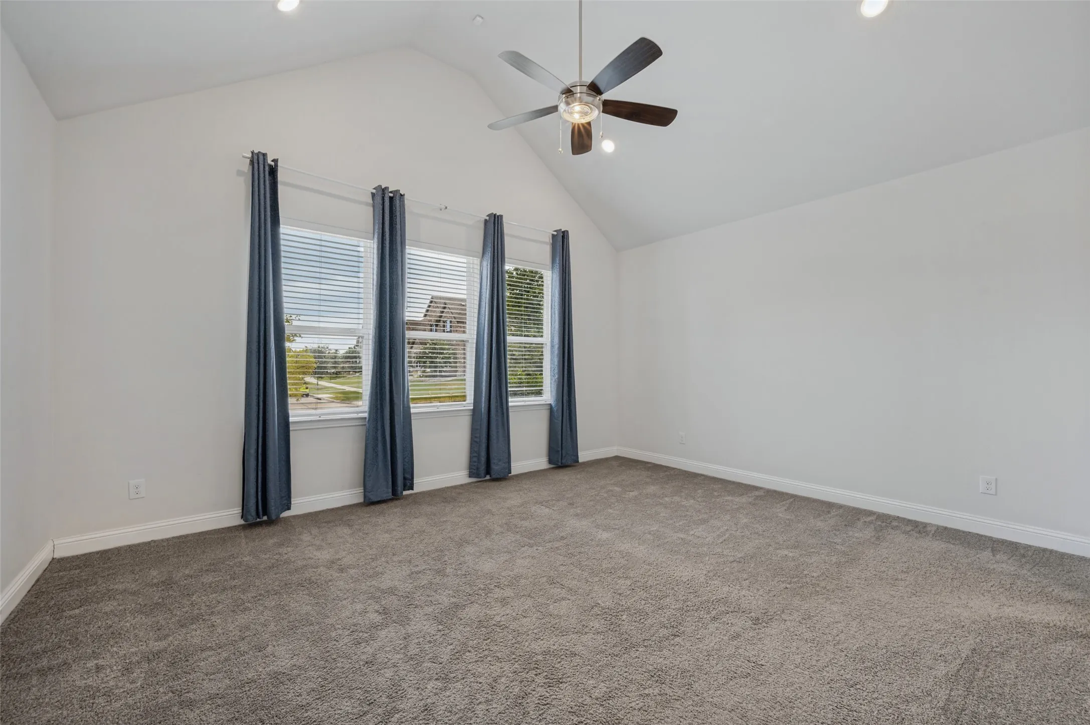Spacious Primary Bedroom with vaulted ceiling, ceiling fan, recessed lighting, plush grey carpeting, and three windows with white blinds.