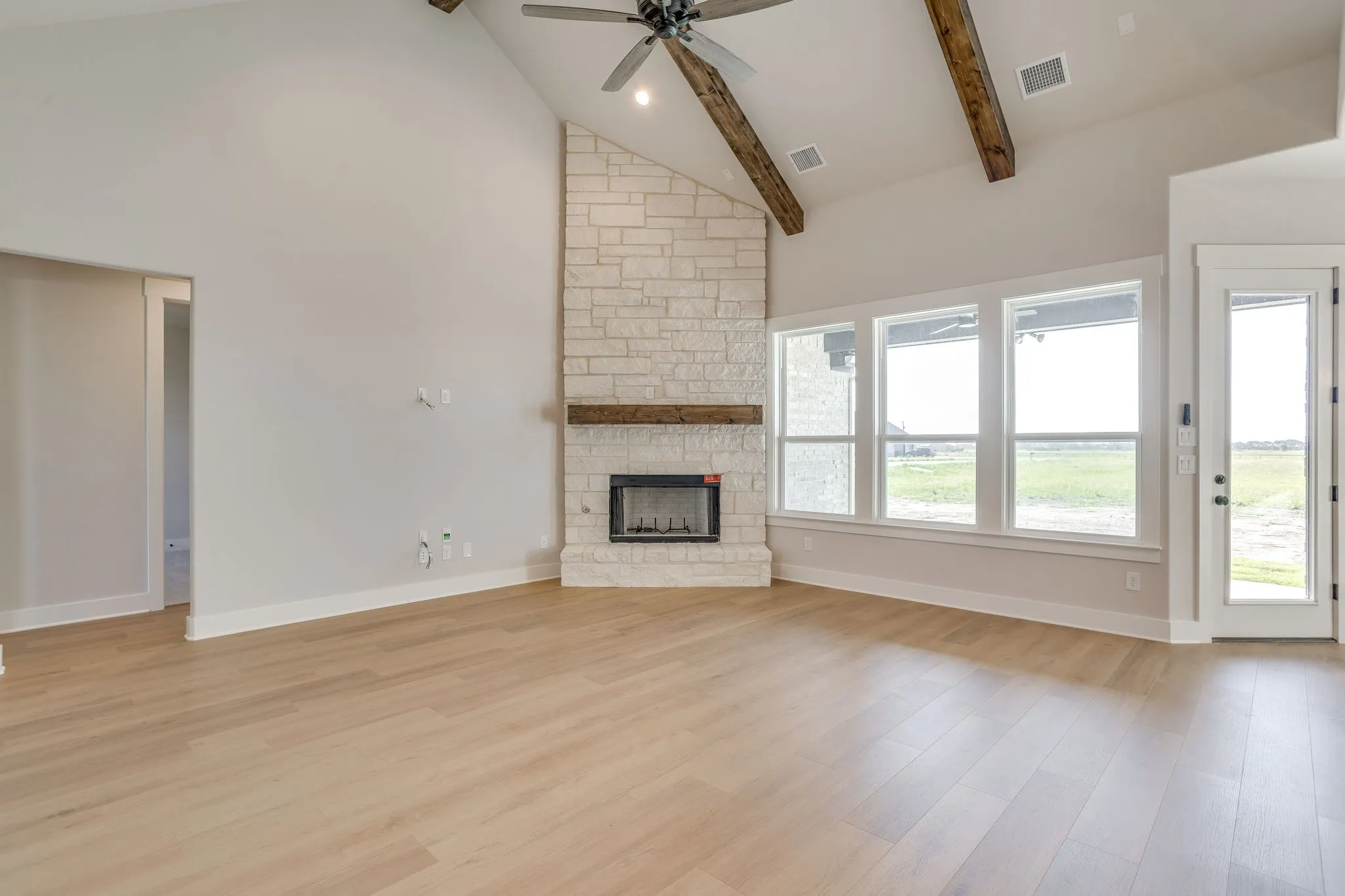 Vaulted ceiling with exposed wood beams, stone fireplace with wood mantel, wood-finish flooring, ceiling fan, and multiple large​​‌​​​​‌​​‌‌​‌‌​​​‌‌​‌​‌​‌​​​‌​​ windows
