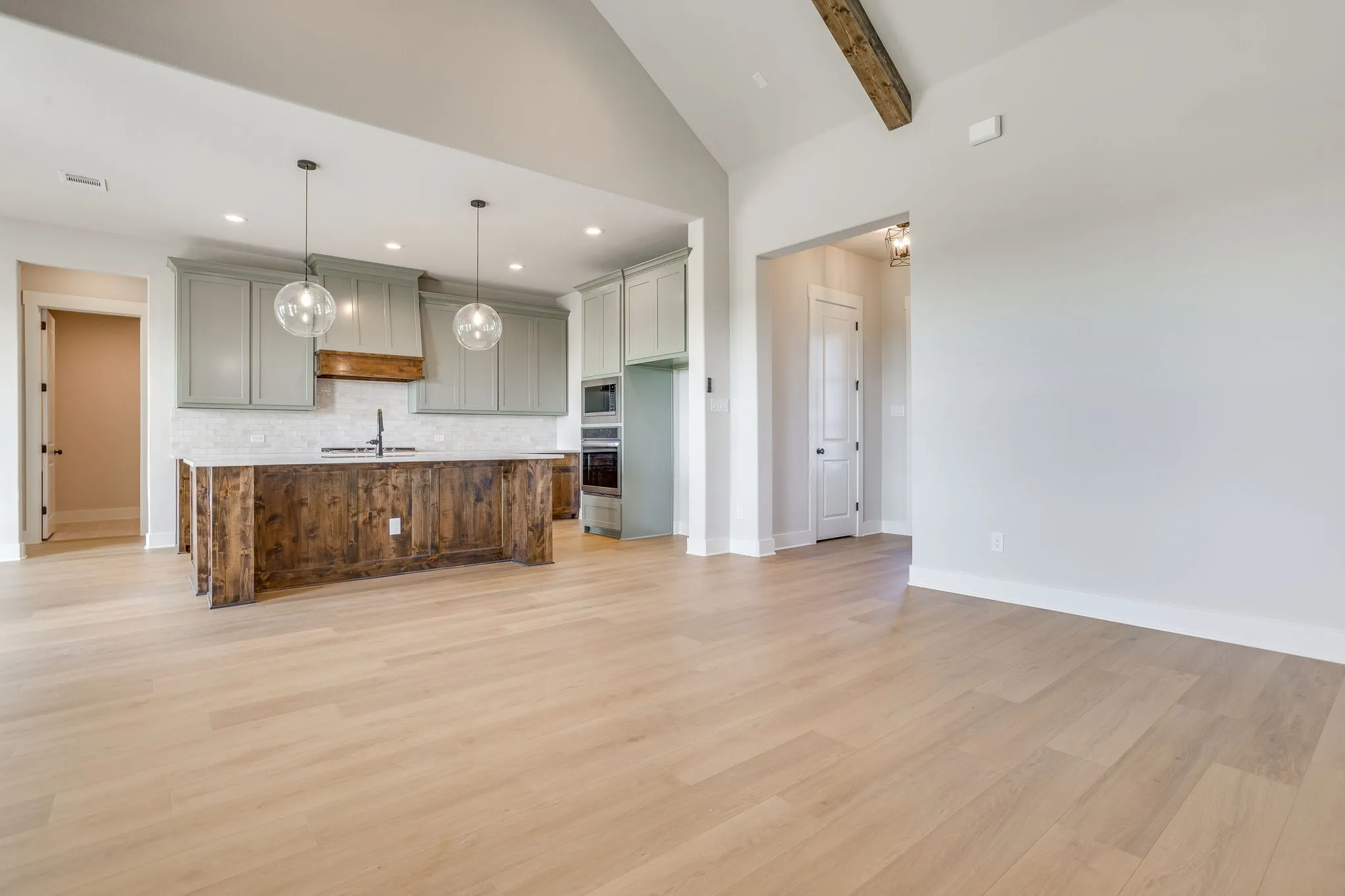 Vaulted ceiling with exposed wood beam, light wood-finish flooring, kitchen island with wood paneling, light green cabinetry, and pendant lighting
