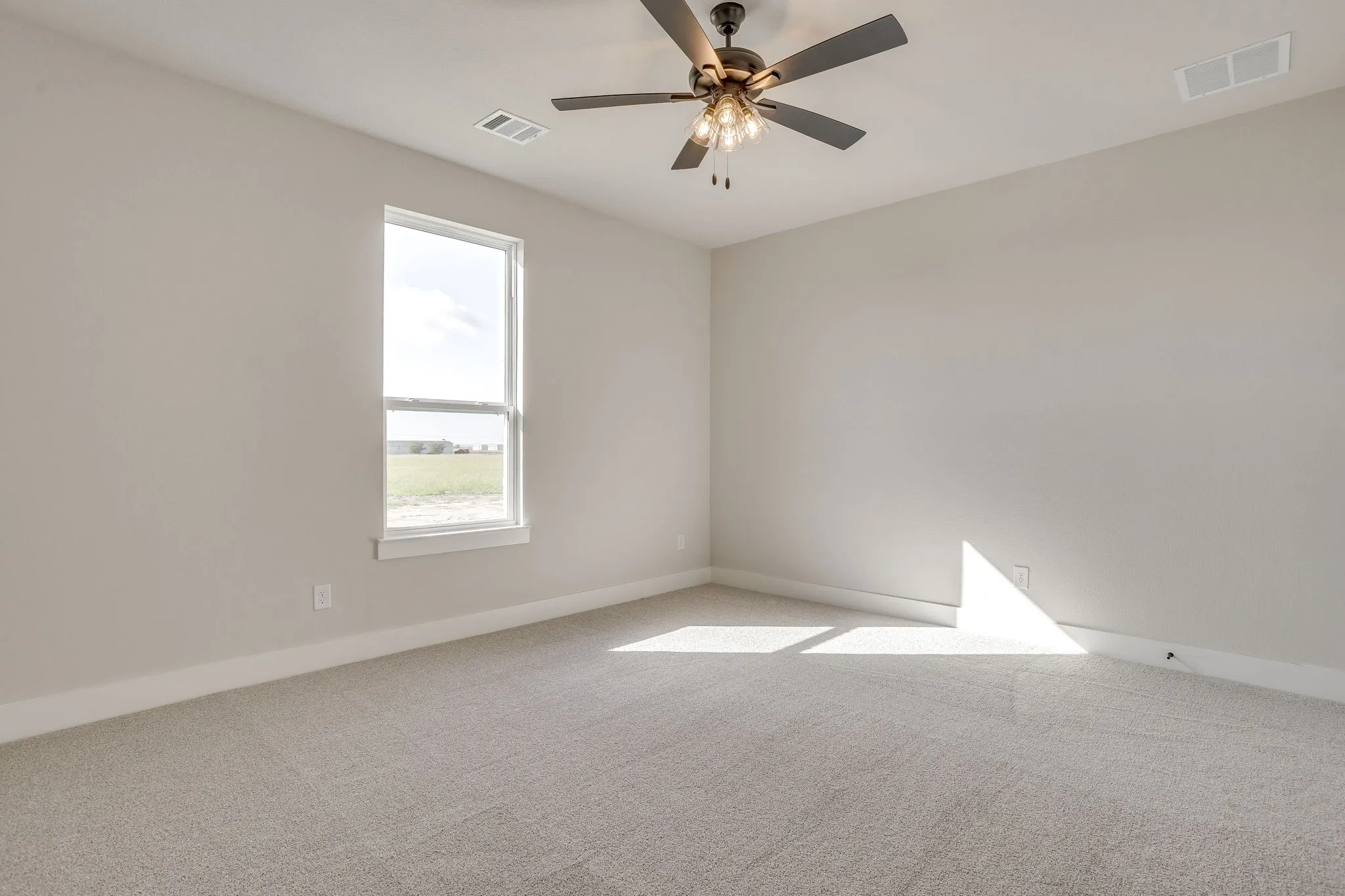 Neutral tone wall paint, light-colored carpeting, a ceiling fan with integrated lighting, and a single window offering exterior scenery