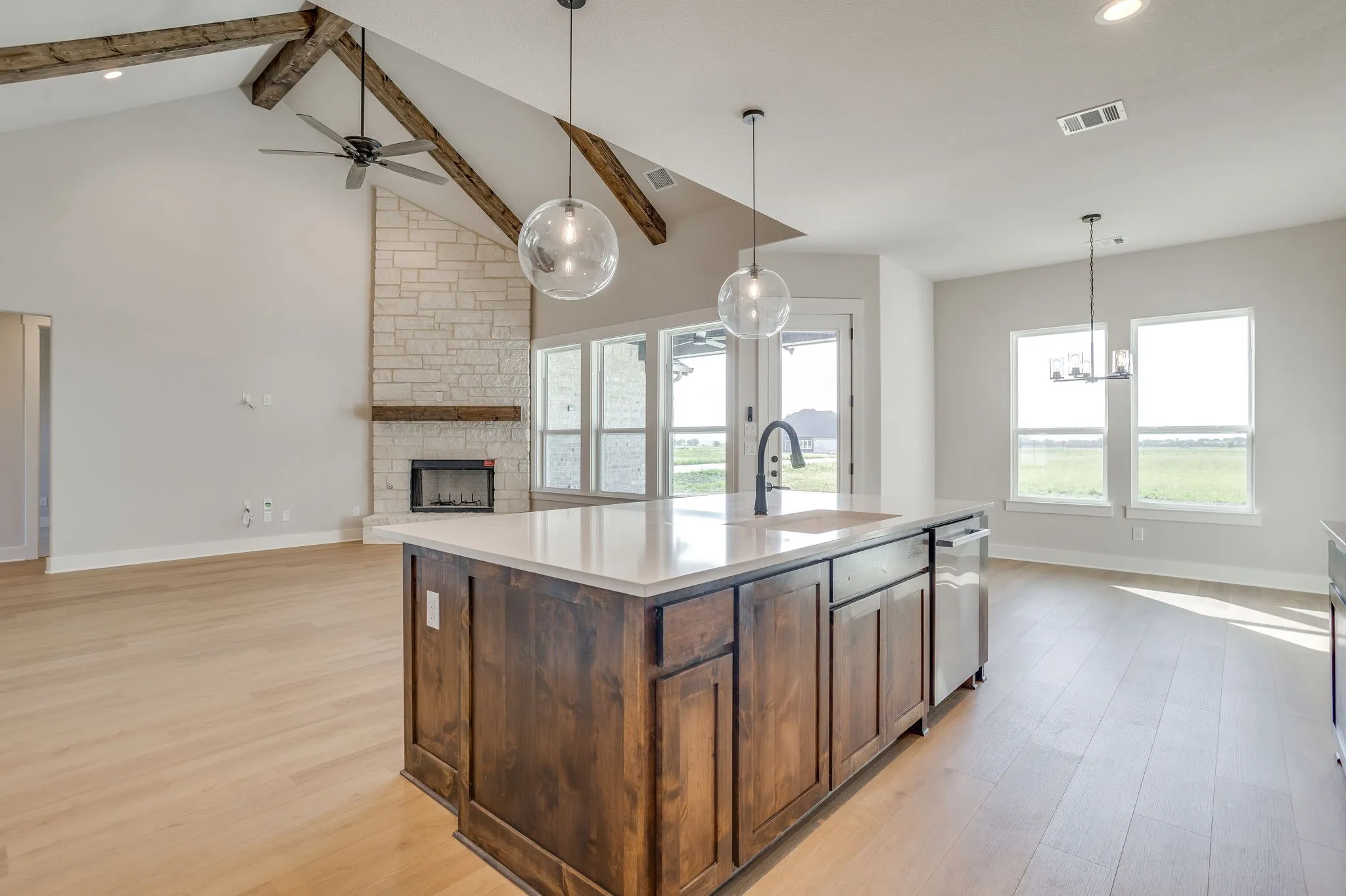 Vaulted ceiling with exposed wood beams, stone fireplace with a wood mantel, central island with an undermount sink and quartz countertop, wood-finish flooring, and pendant lighting