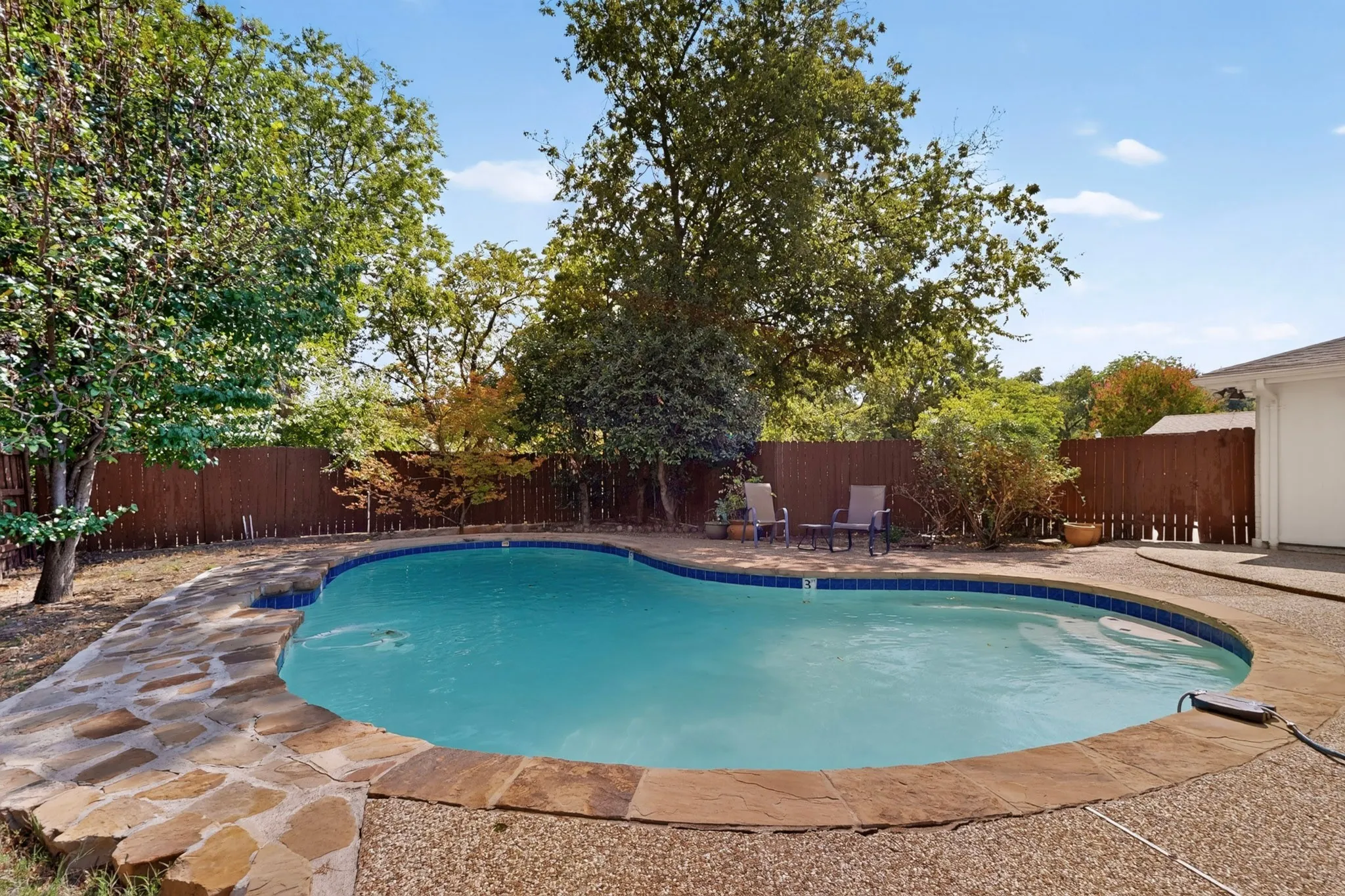 Curved swimming pool with stone coping, surrounded by a flagstone and aggregate concrete patio, and a privacy fence