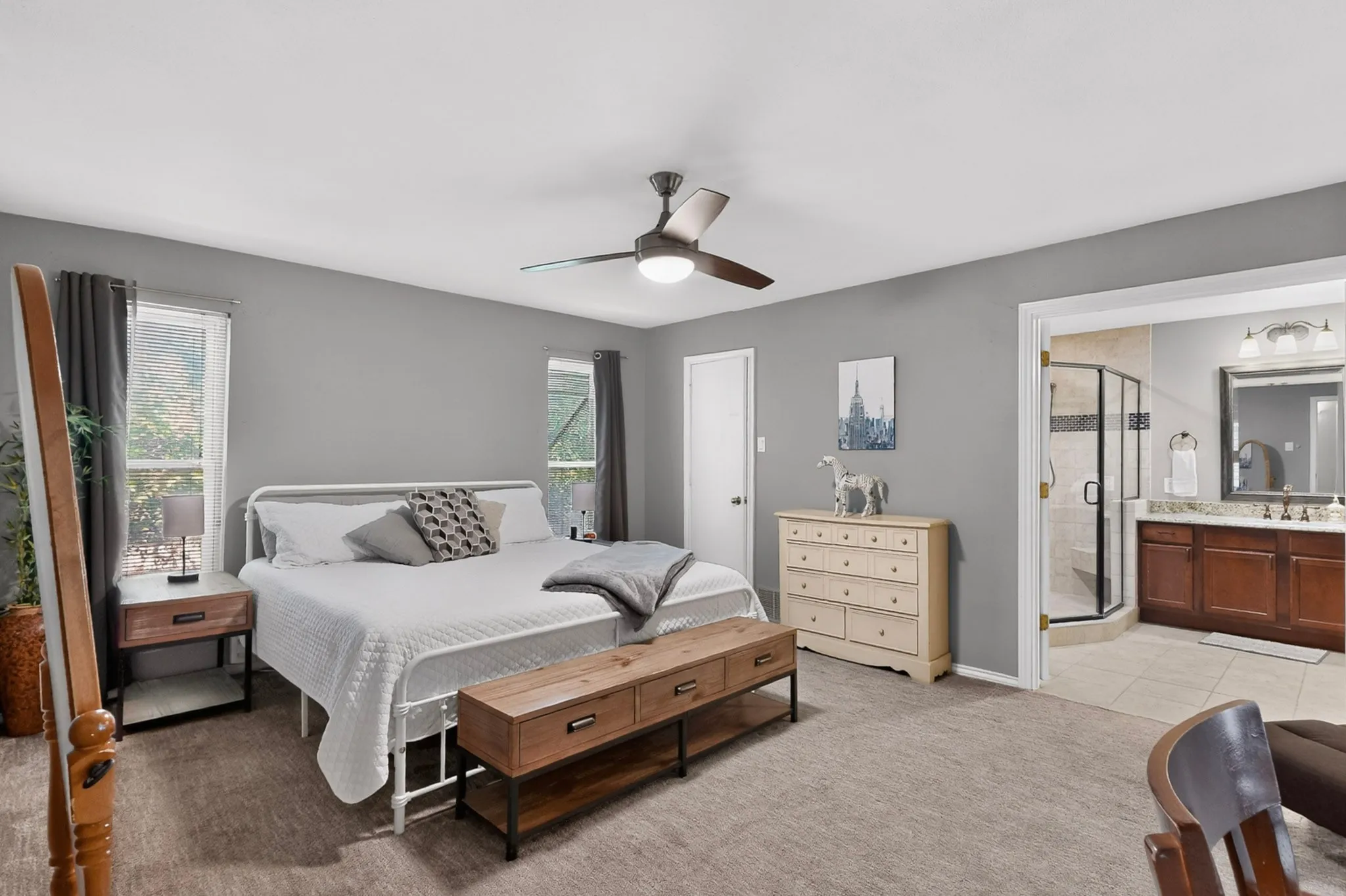 Spacious bedroom featuring neutral wall tones, two windows with dark drapery, a ceiling fan, and direct access to an ensuite bathroom
