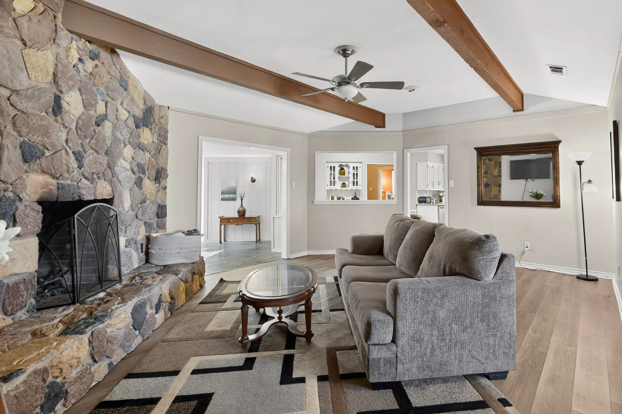 Stone-clad fireplace with hearth, wood-finish flooring, exposed ceiling beams, and a ceiling fan