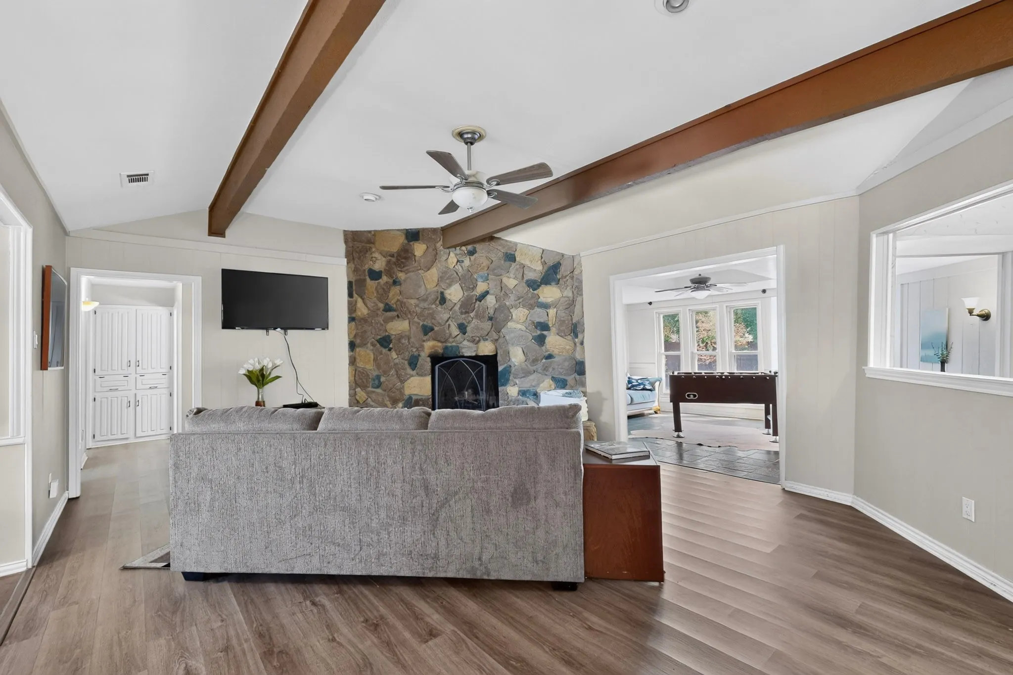 Vaulted ceiling with wood beams, stone-faced fireplace, wood-finish flooring, and a ceiling fan
