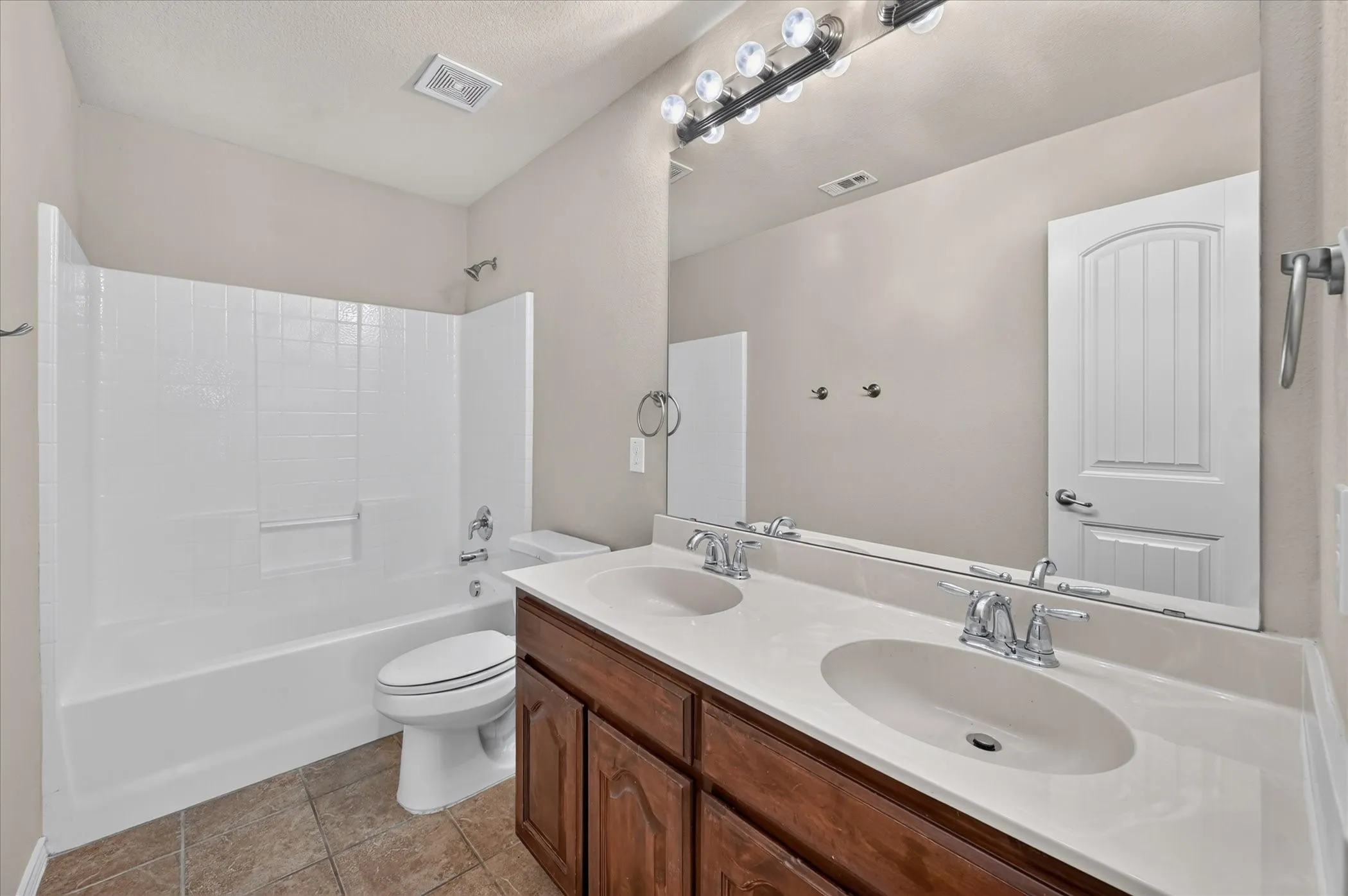 Dual basin vanity with wood-finish cabinetry, broad wall mirror, and tile flooring