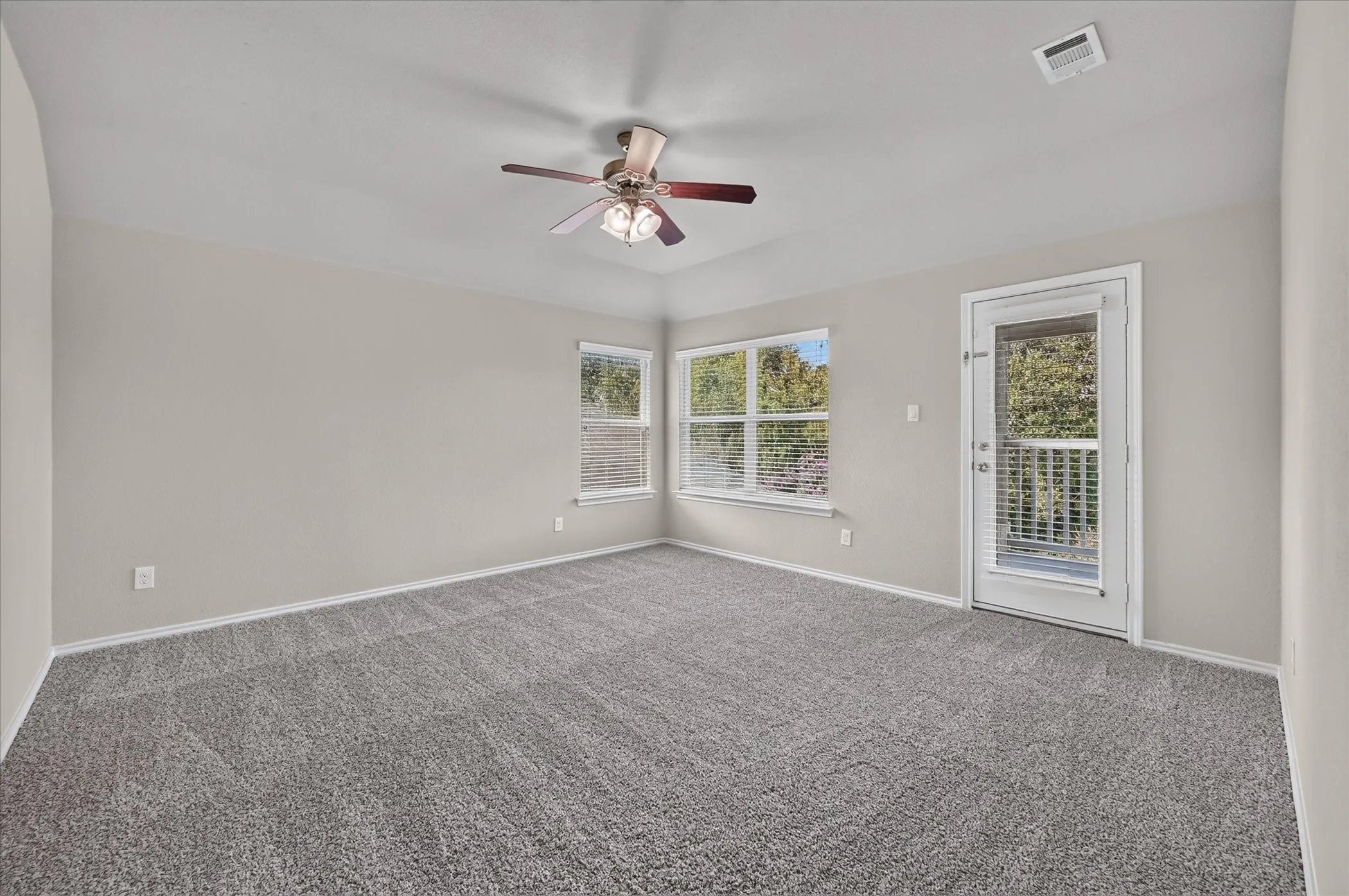 Neutral-toned wall paint throughout, plush gray carpeting, ceiling fan with integrated lighting, multiple windows with blinds, and a glass-paneled door leading to a balcony