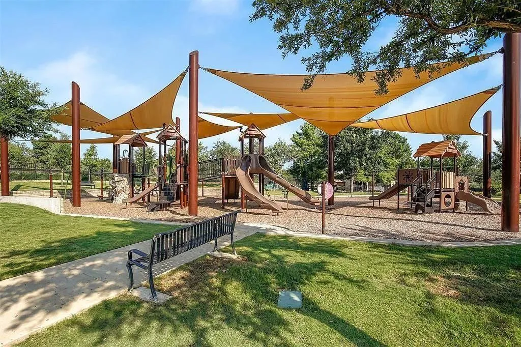 Expansive playground featuring multiple play structures, overhead shade sails, a paved pathway, and surrounding grassy areas