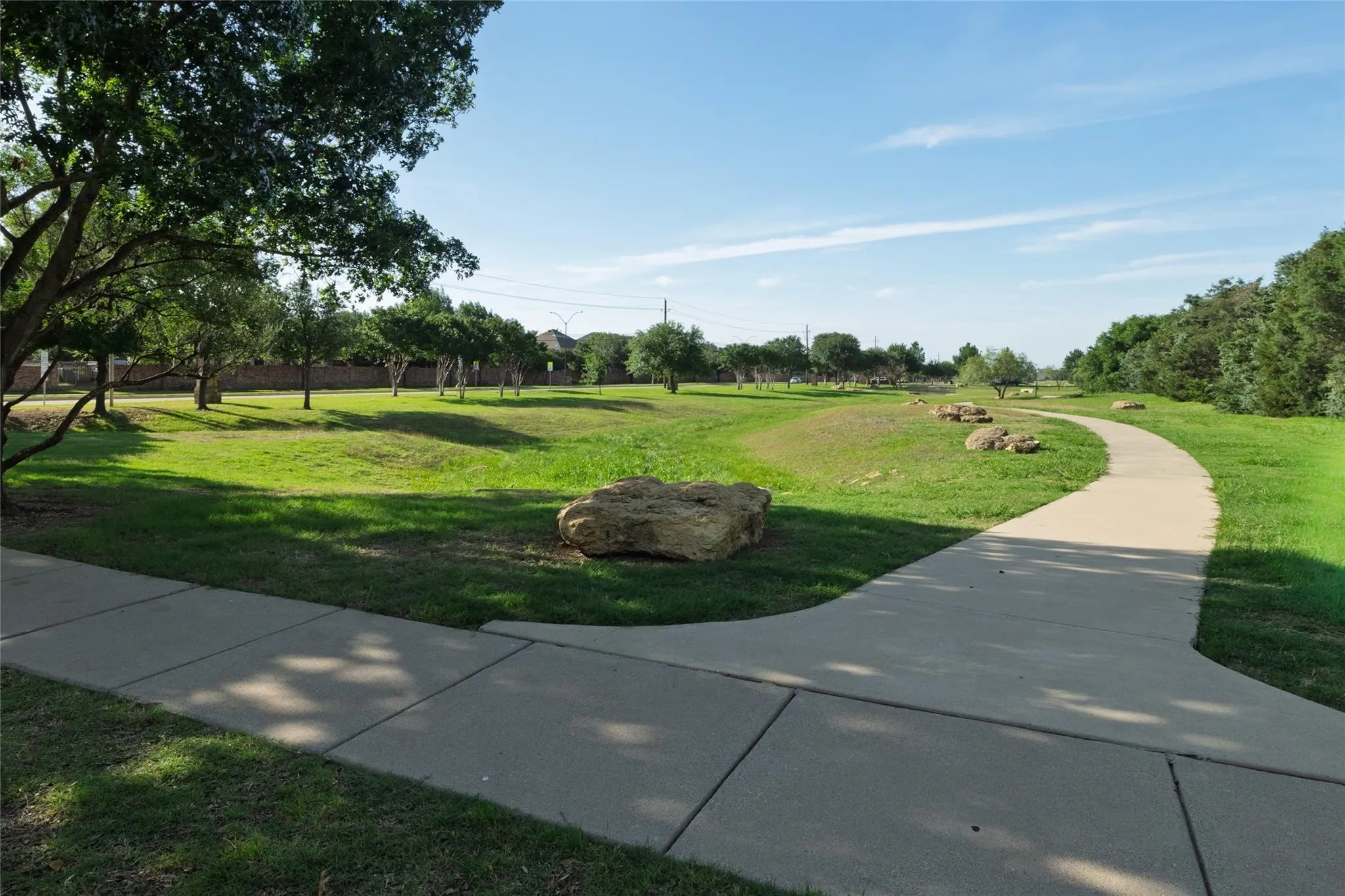 Expansive green space featuring a paved walking path, mature trees, and natural rock accents