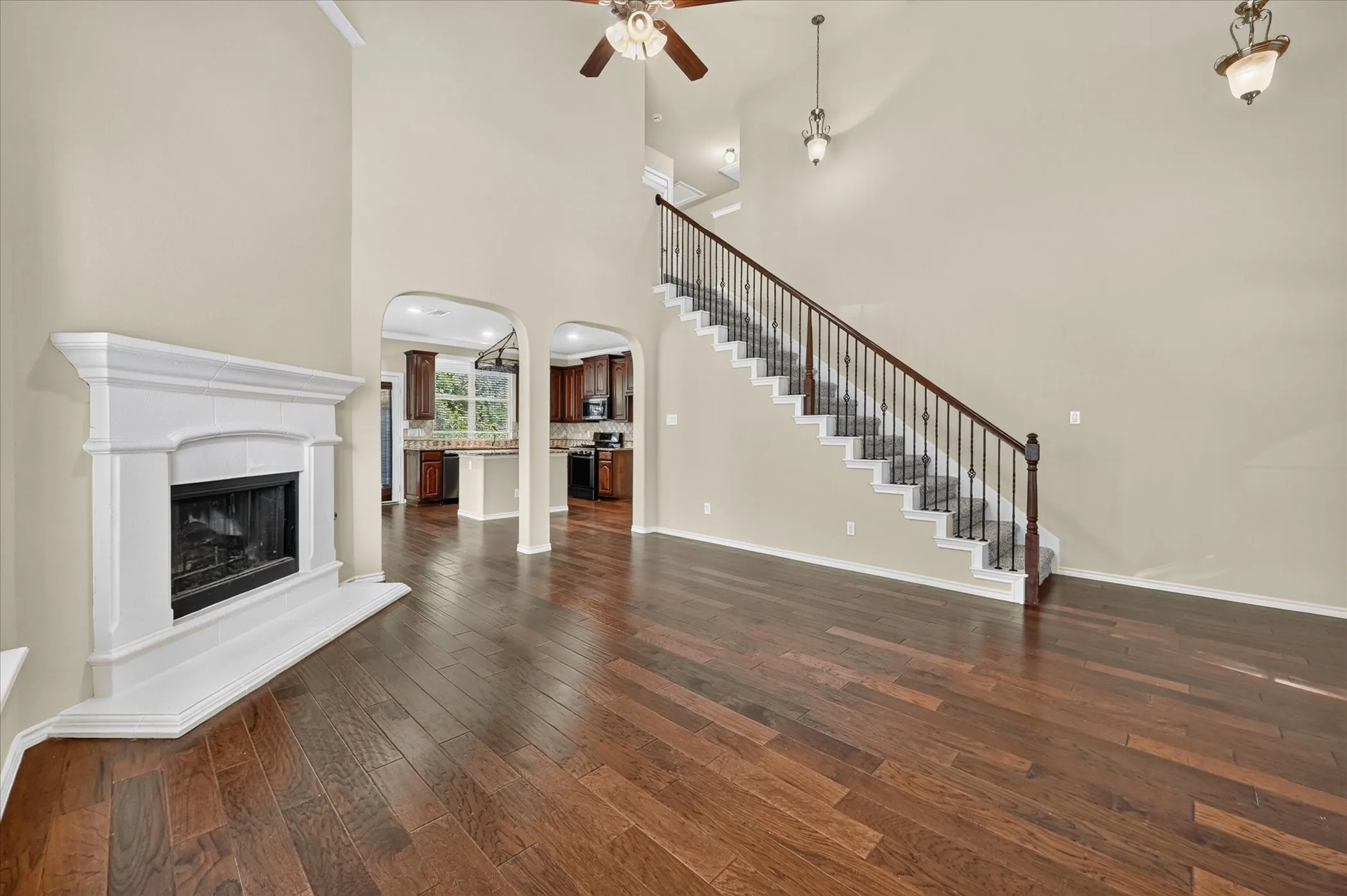 Wood-finish flooring throughout, a white fireplace with a mantel, a staircase with dark wood railing and iron balusters, arched doorways, and a ceiling fan with integrated lighting