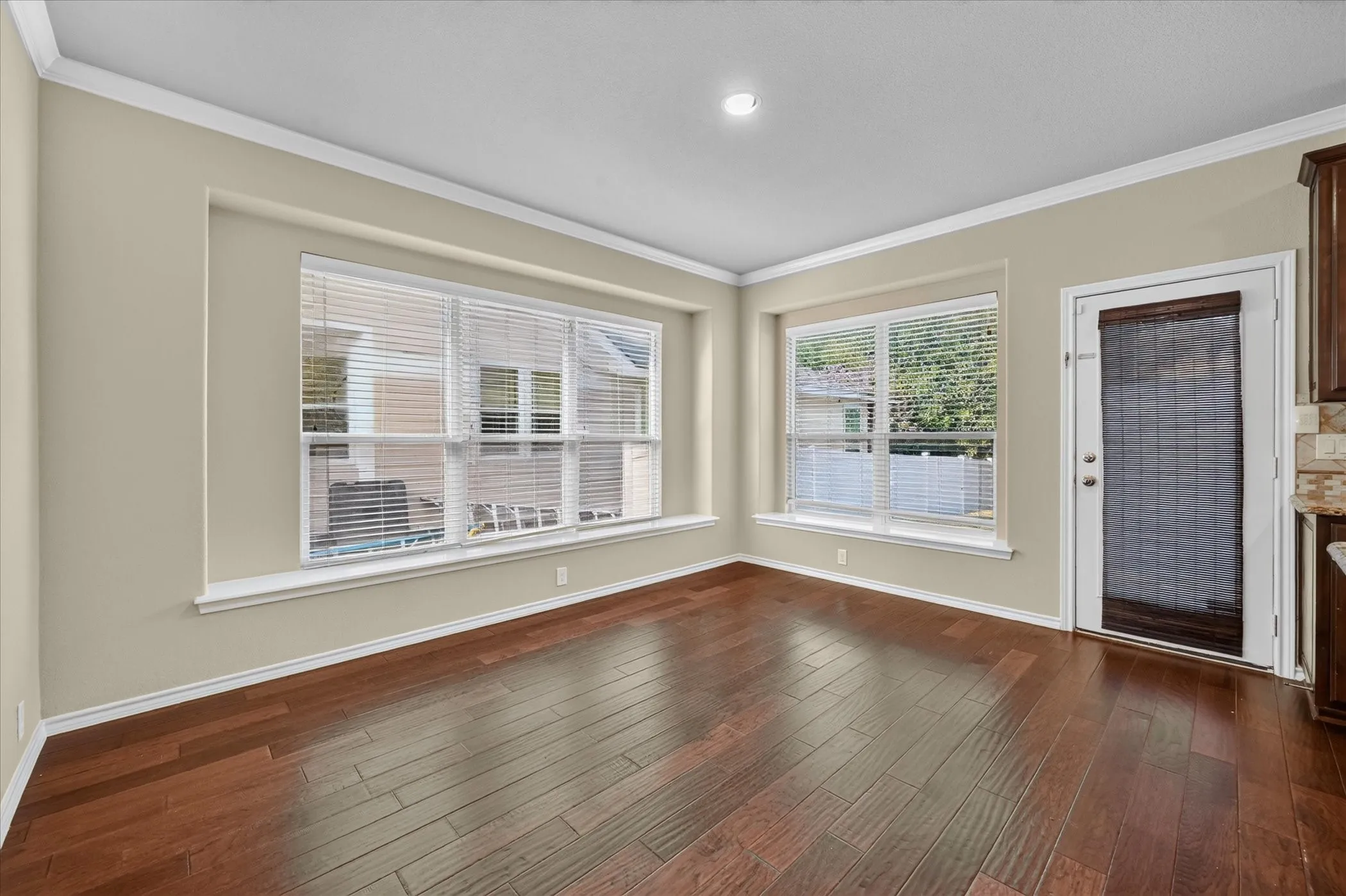 Rich wood-finish flooring, two large windows with white blinds, and a white exterior door with a woven shade
