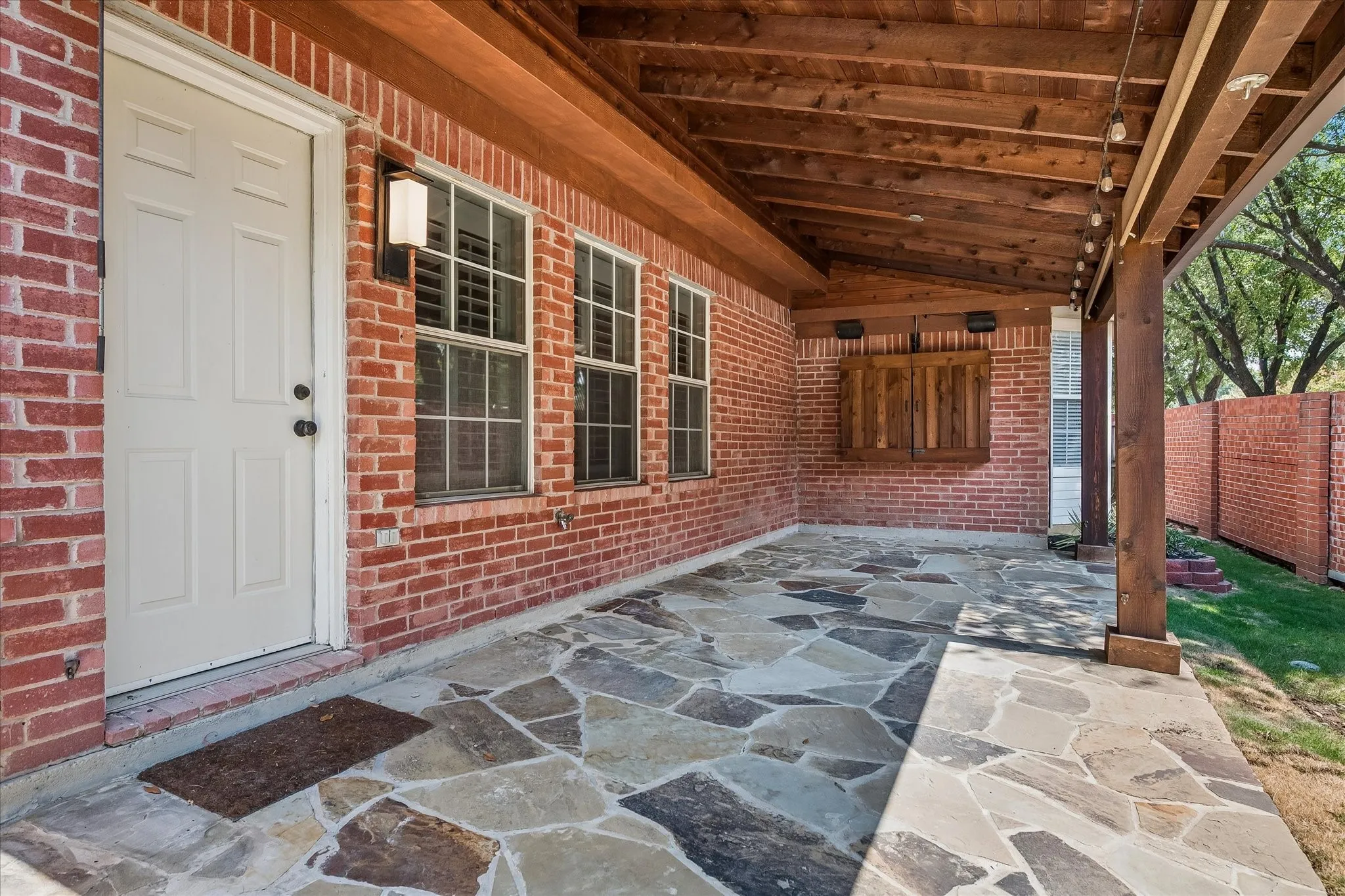 Covered patio with a flagstone floor, exposed wood beam ceiling, brick exterior, and wall-mounted light fixture