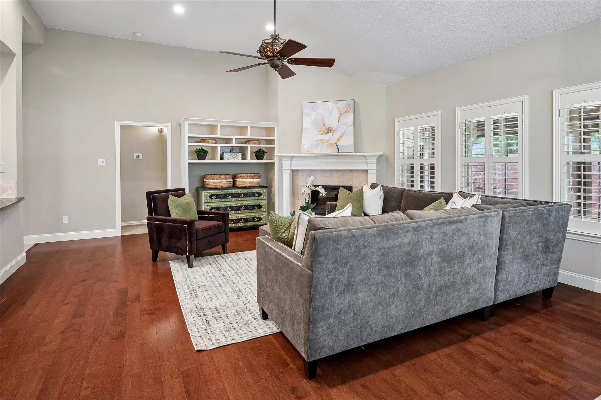 Vaulted ceiling with a decorative ceiling fan, wood-finish flooring, built-in shelving, stone fireplace, and plantation shutters