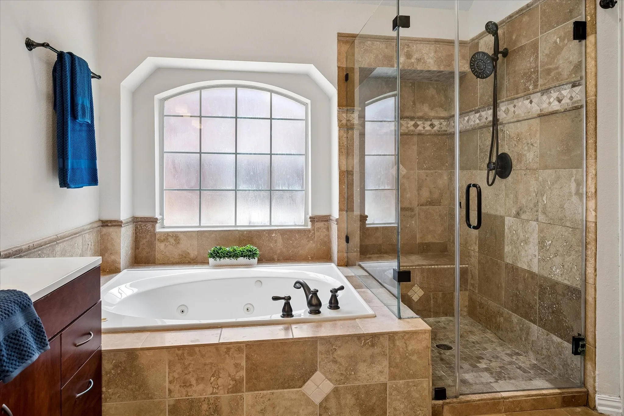 Soaking tub with bronze fixtures, arched window, glass-enclosed shower, tiled shower surround with decorative accent, and built-in shower bench
