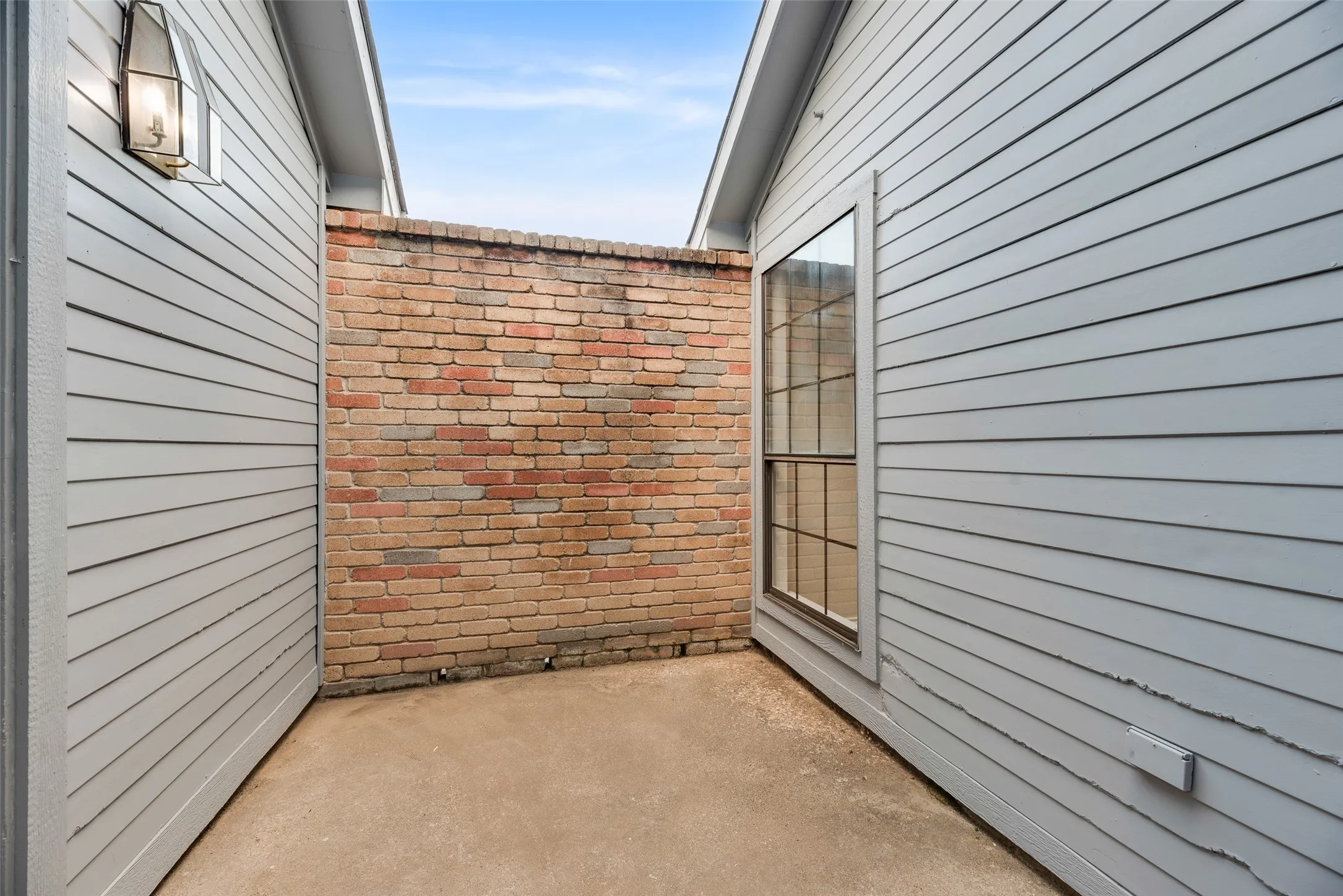 Paved courtyard featuring a brick privacy wall, exterior wall lantern, and a multi-pane window