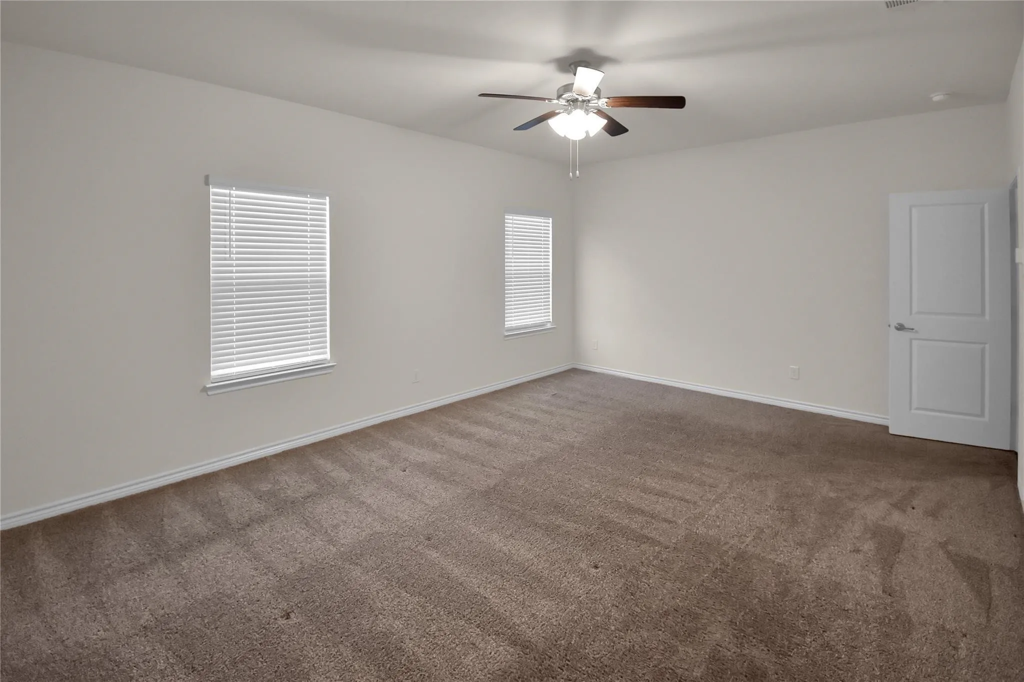 Spacious room featuring neutral carpet, two windows with blinds, a ceiling fan, and a white paneled door