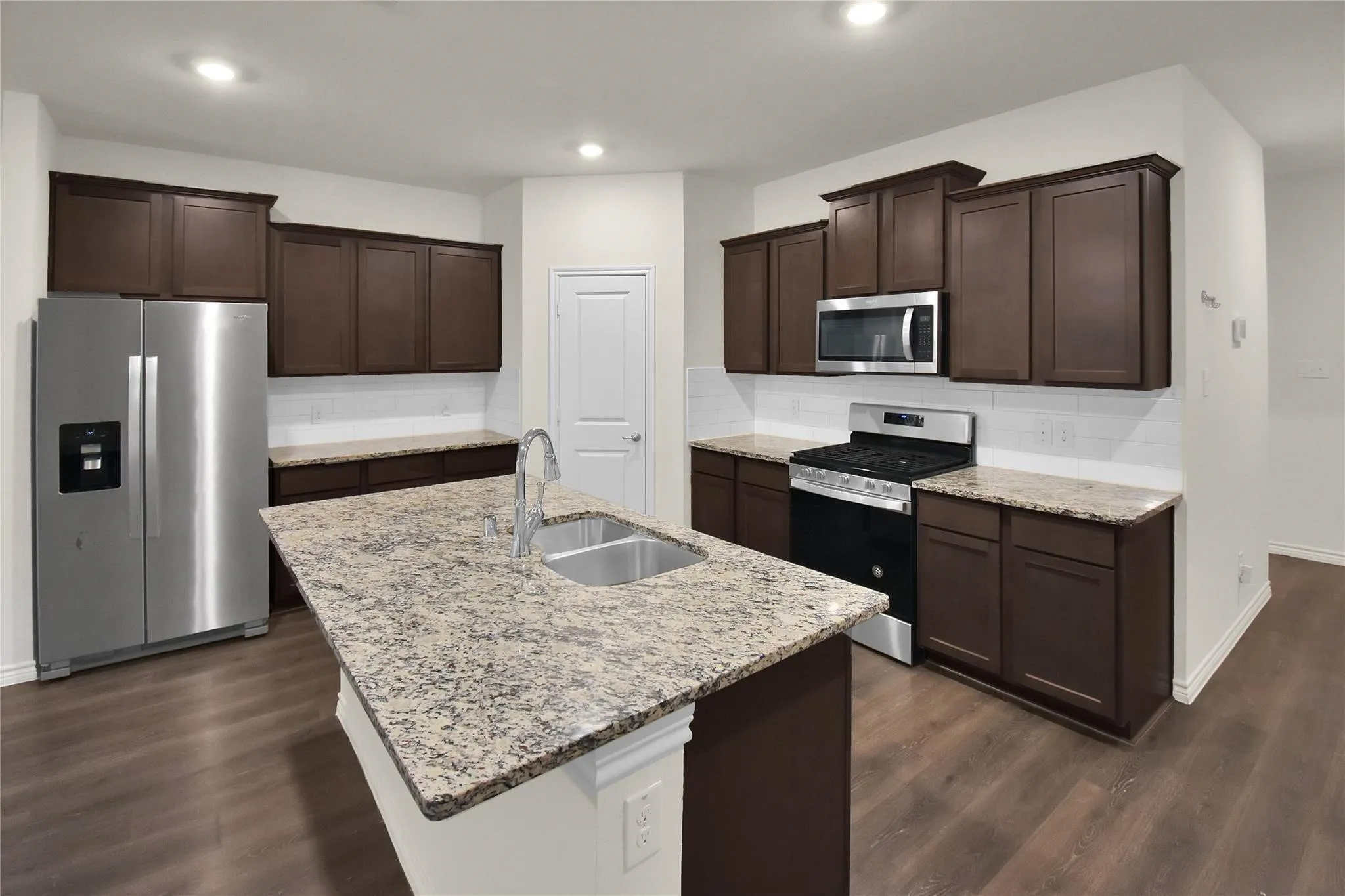 Kitchen featuring an island with an undermount sink, dark wood-finish cabinetry, stainless steel appliances, a tile backsplash, and light-toned countertops