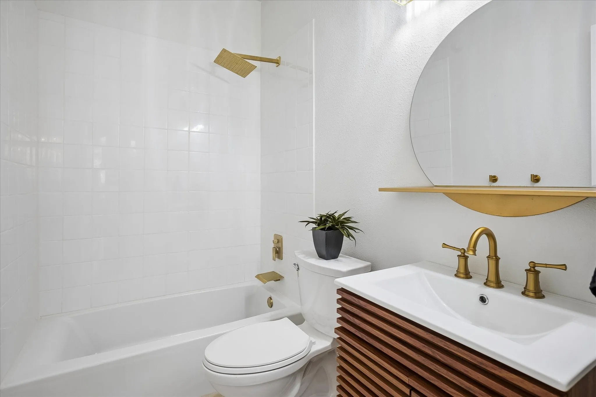 Full white subway tile bath surround, gold-finish rain shower head, round wall-mounted mirror, wood-finish vanity, and gold-finish faucet