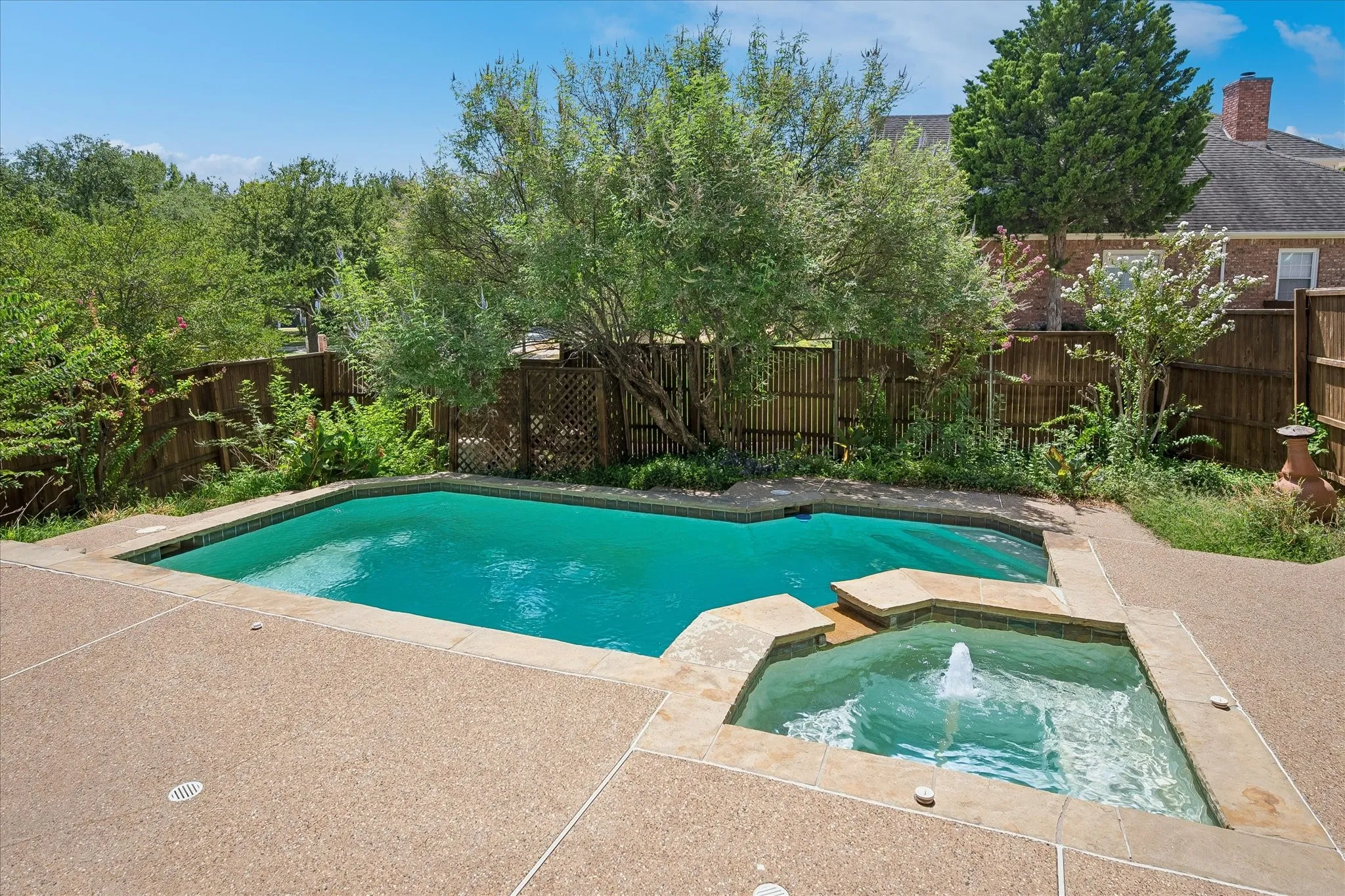 Swimming pool with a spa, surrounded by a textured concrete patio and mature trees, featuring a wooden fence
