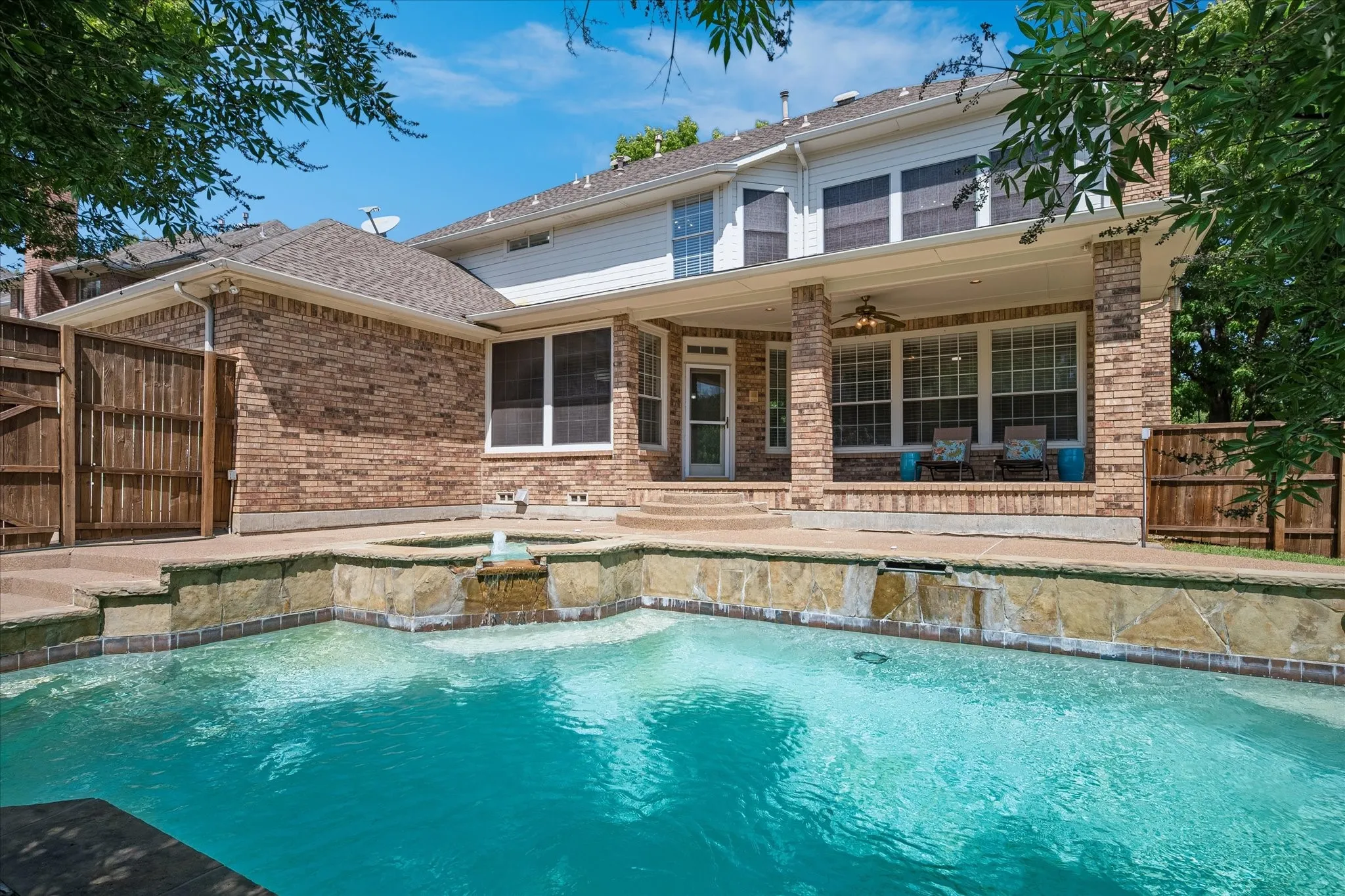 Outdoor swimming pool with integrated spa and waterfall feature, expansive covered patio with brick columns, and a two-story brick and siding exterior