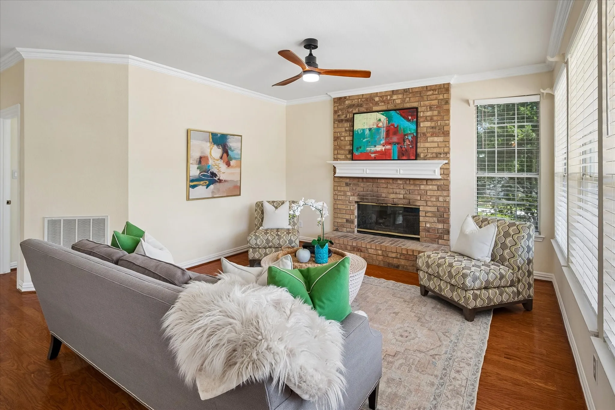 Brick fireplace with a white mantel, wood-finish flooring, white crown molding, a ceiling fan with light, and multiple windows