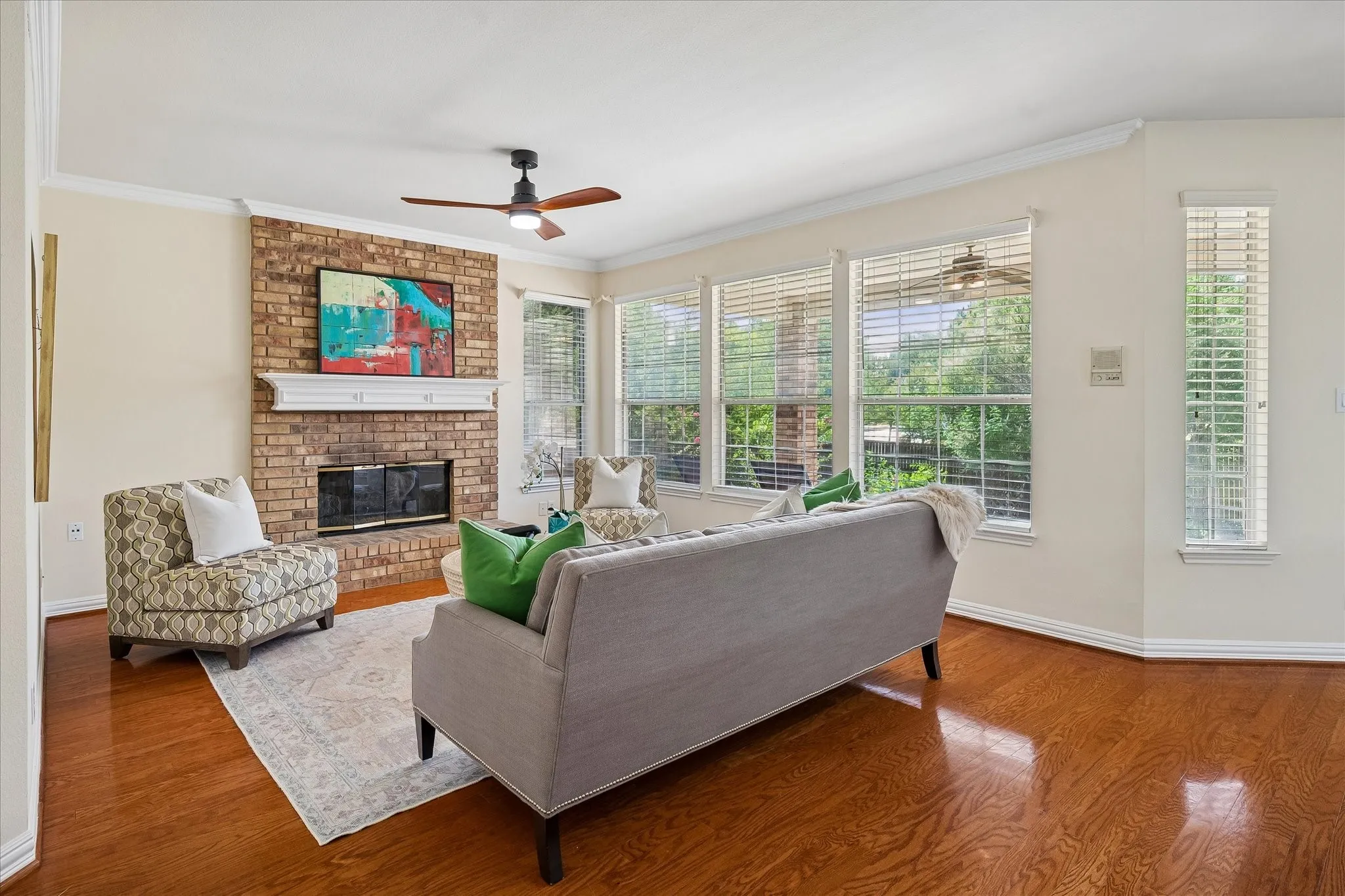 Hardwood-finish flooring, brick-faced fireplace with white mantel, ceiling fan with integrated lighting, and multiple windows with blinds