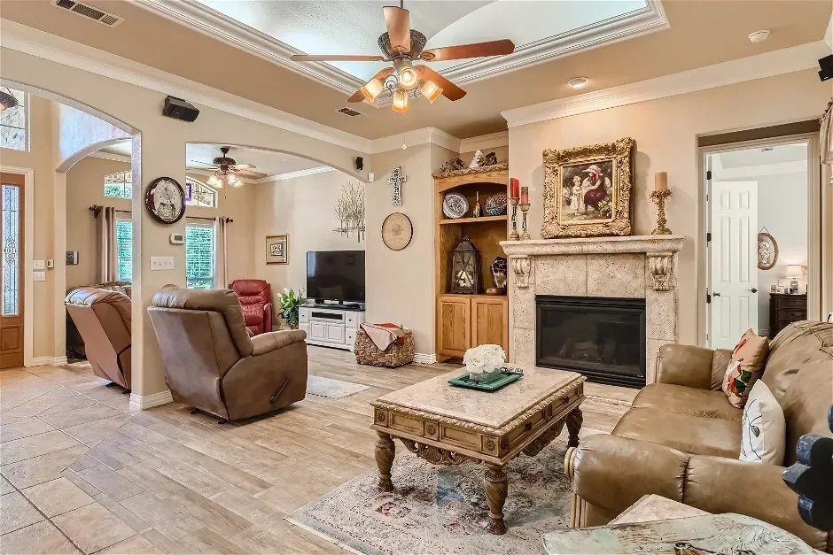 Tray ceiling with a ceiling fan, stone-surround fireplace with a mantel, built-in shelving, wood-finish flooring, and arched doorways