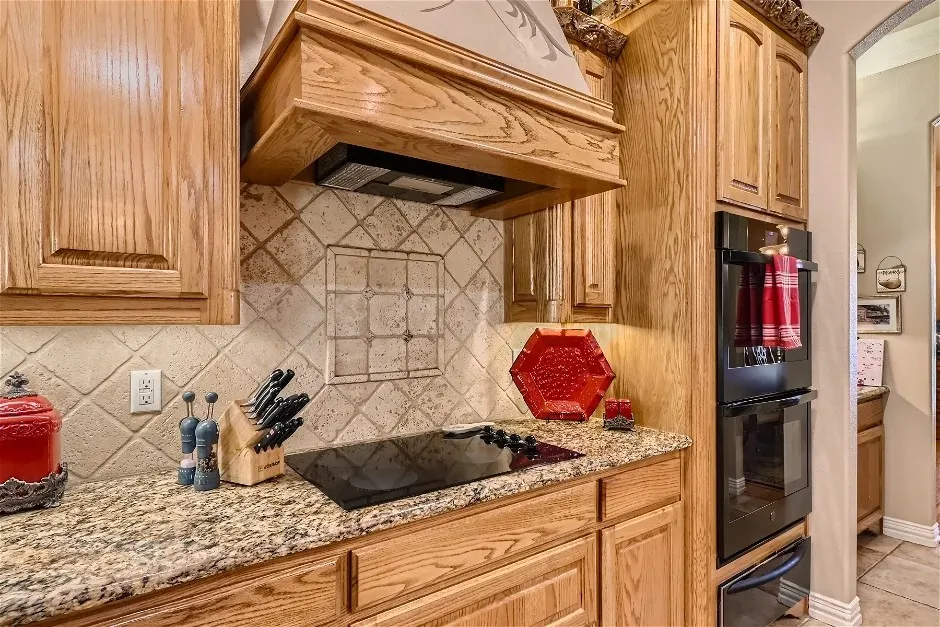 Wood cabinetry with decorative crown molding, stone countertops, tiled backsplash with decorative inlay, built-in cooktop, and double wall ovens