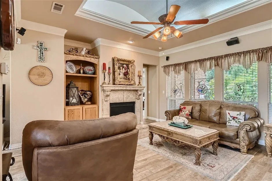 Tray ceiling with crown molding, built-in shelving, fireplace with decorative mantel, ceiling fan with light fixture, and wood-finish flooring